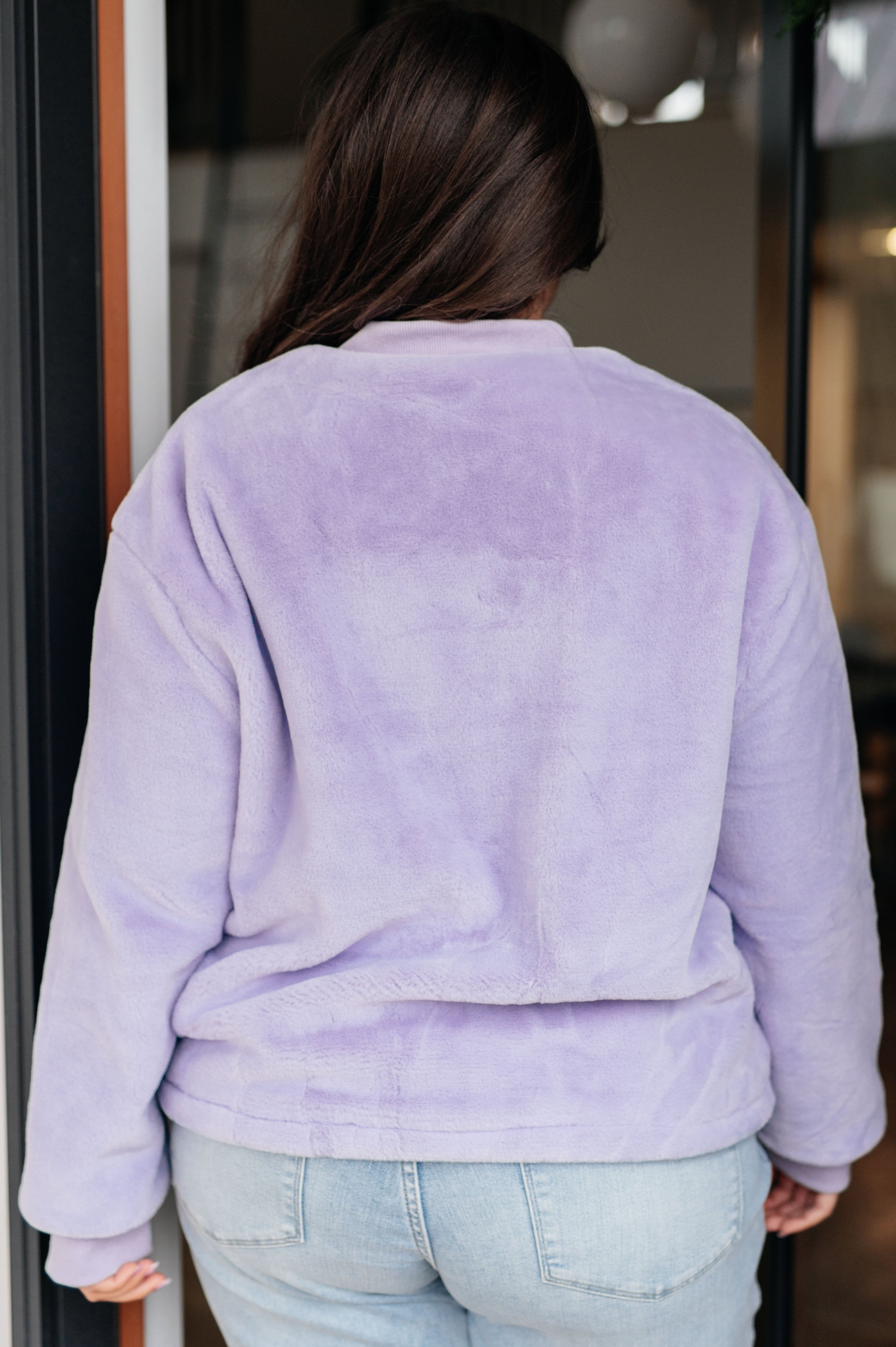 Woman wearing a light purple fuzzy zip up cropped coat and light blue jeans standing in front of a glass door.