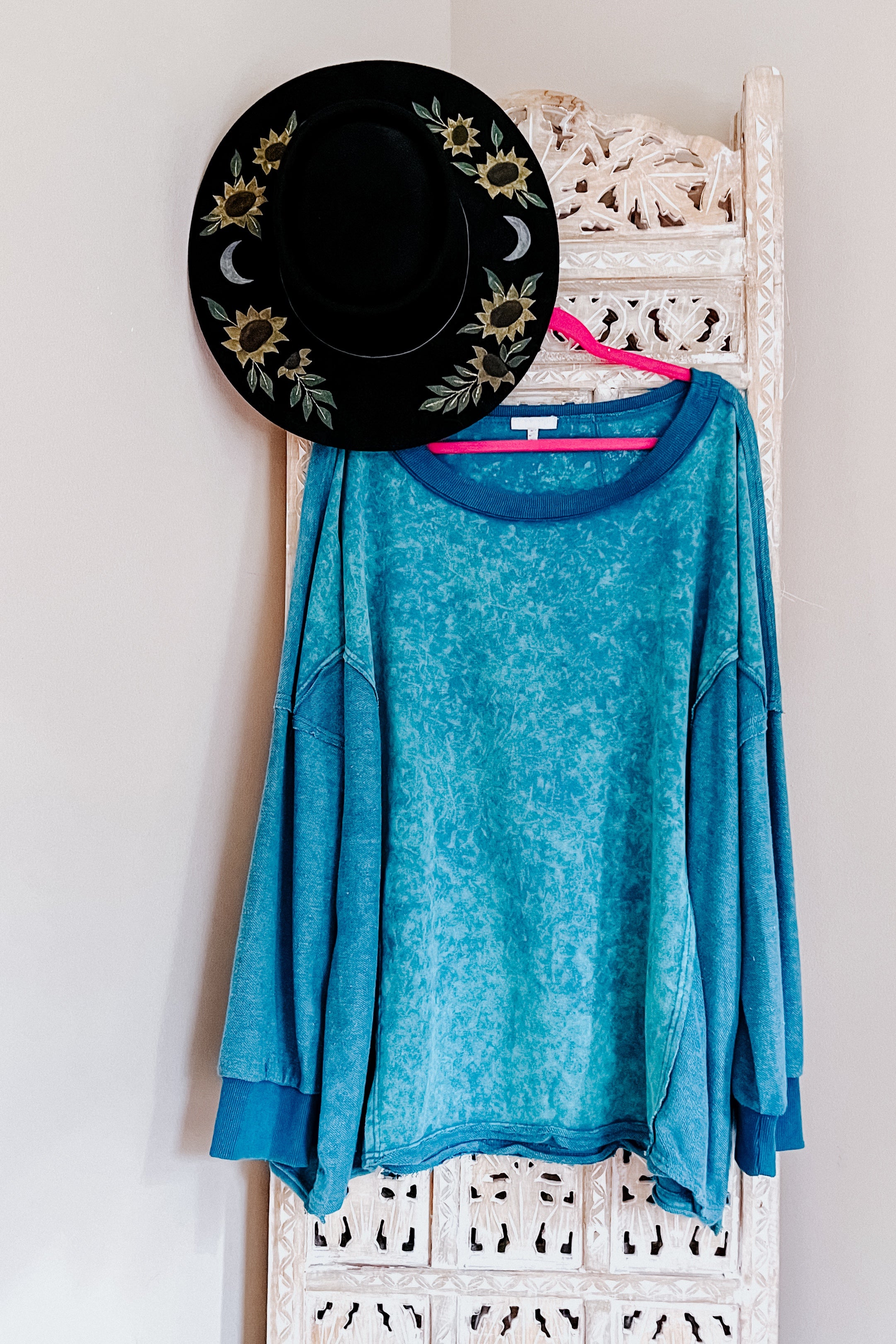 A blue mineral wash pullover on a pink hanger hanging on a rustic room divider against a white wall with a black sunflower hat.