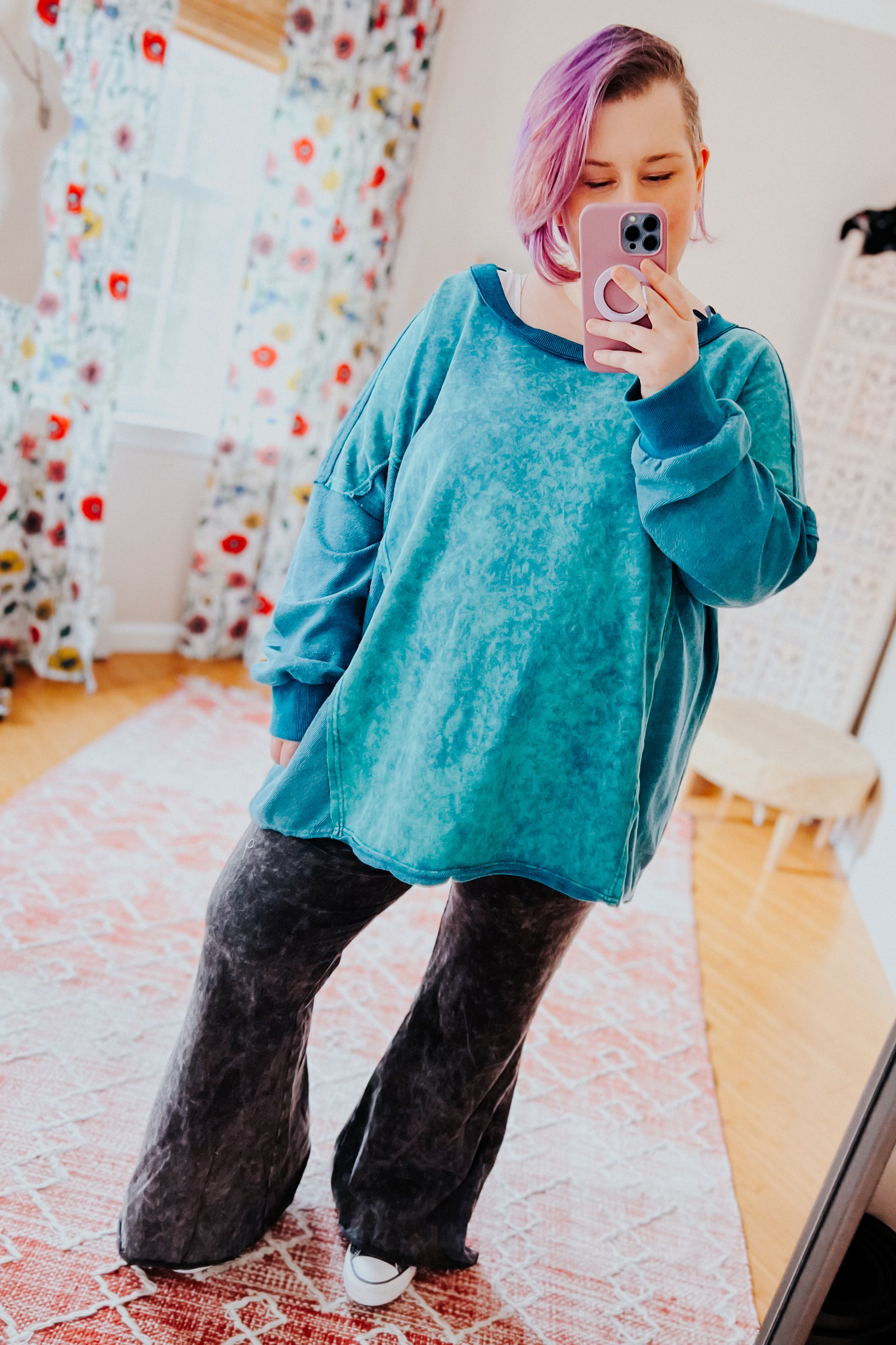 A woman with pink hair wearing a blue mineral wash sweater over dark grey flare pants against an indoor setting.