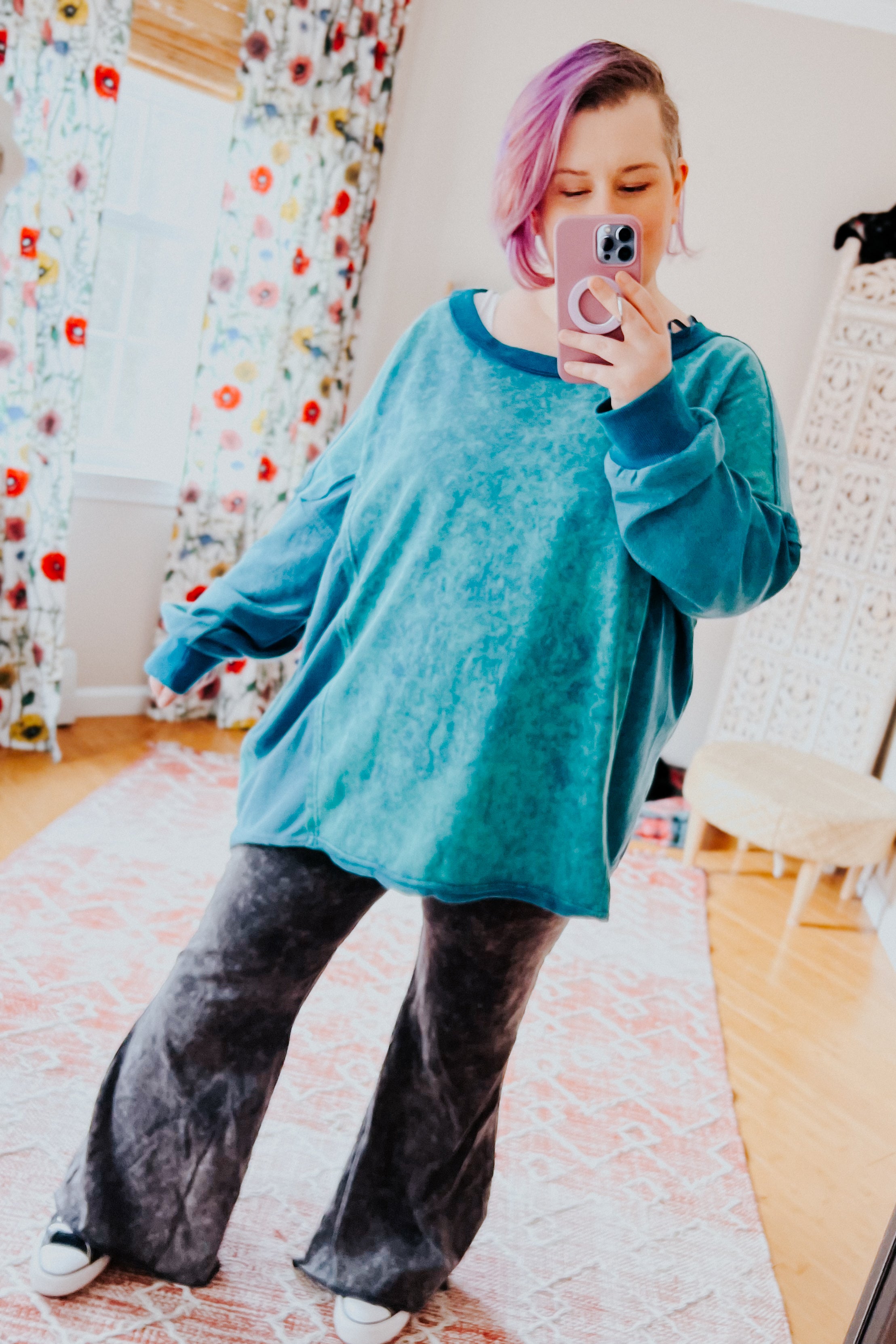 A woman with pink hair wearing a blue mineral wash sweater over dark grey flare pants against an indoor setting.