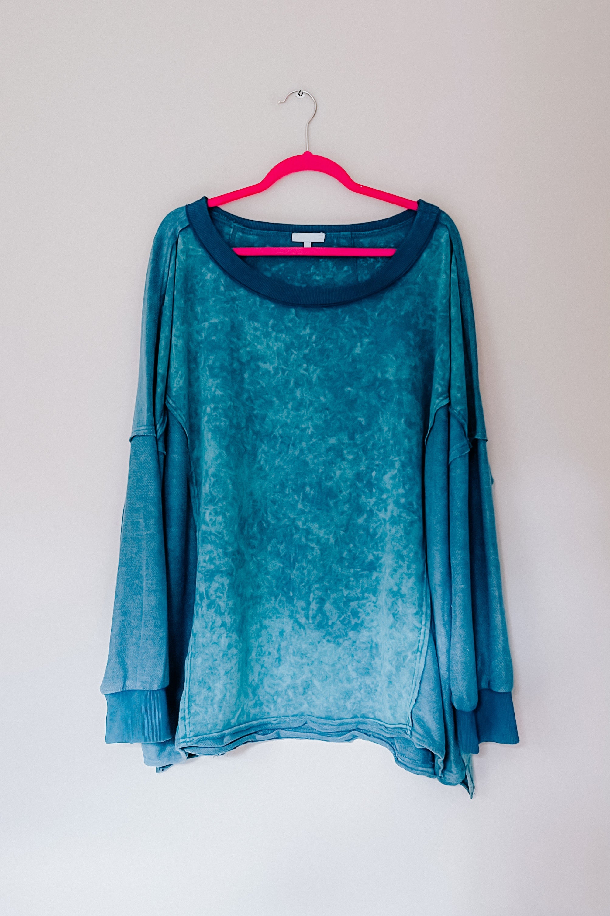 A blue mineral wash pullover handing on a pink hanger against a white backdrop.