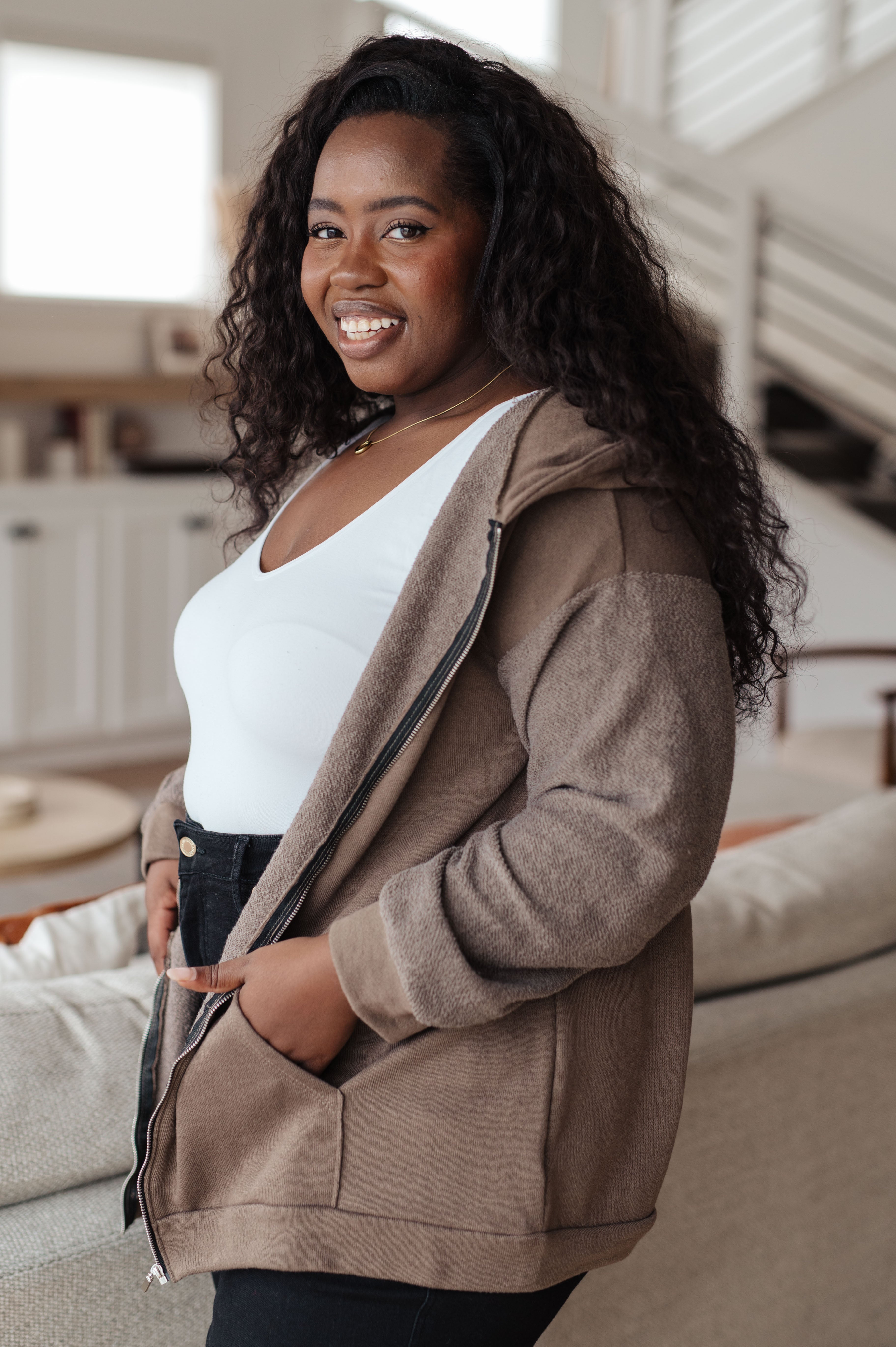 Woman wearing a brown zip-up hoodie over a white tank top over black jeans in a casual indoor setting.