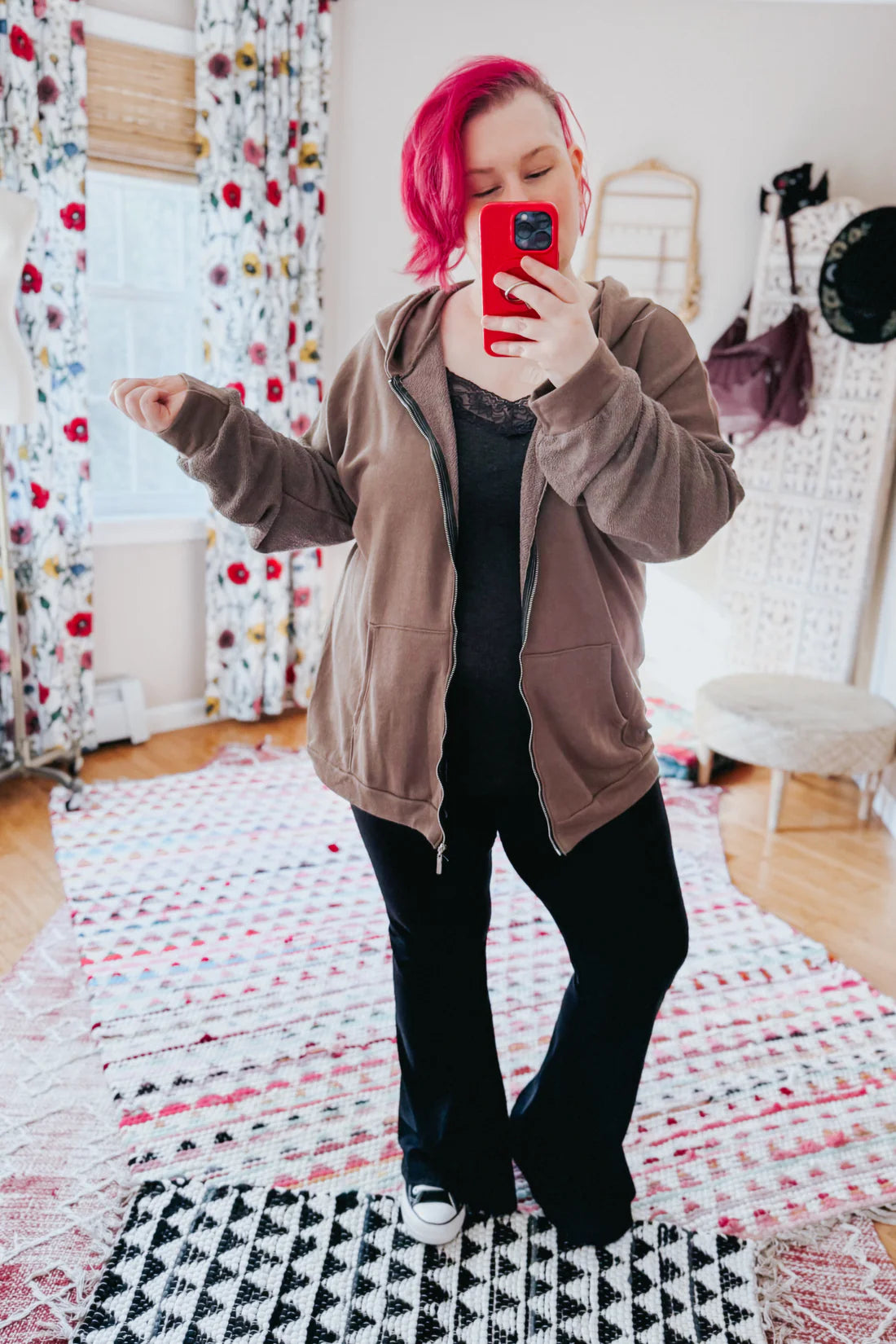 A woman with red hair wearing a brown zip up hoodie, a grey lace top and black flare pants against an indoor setting.