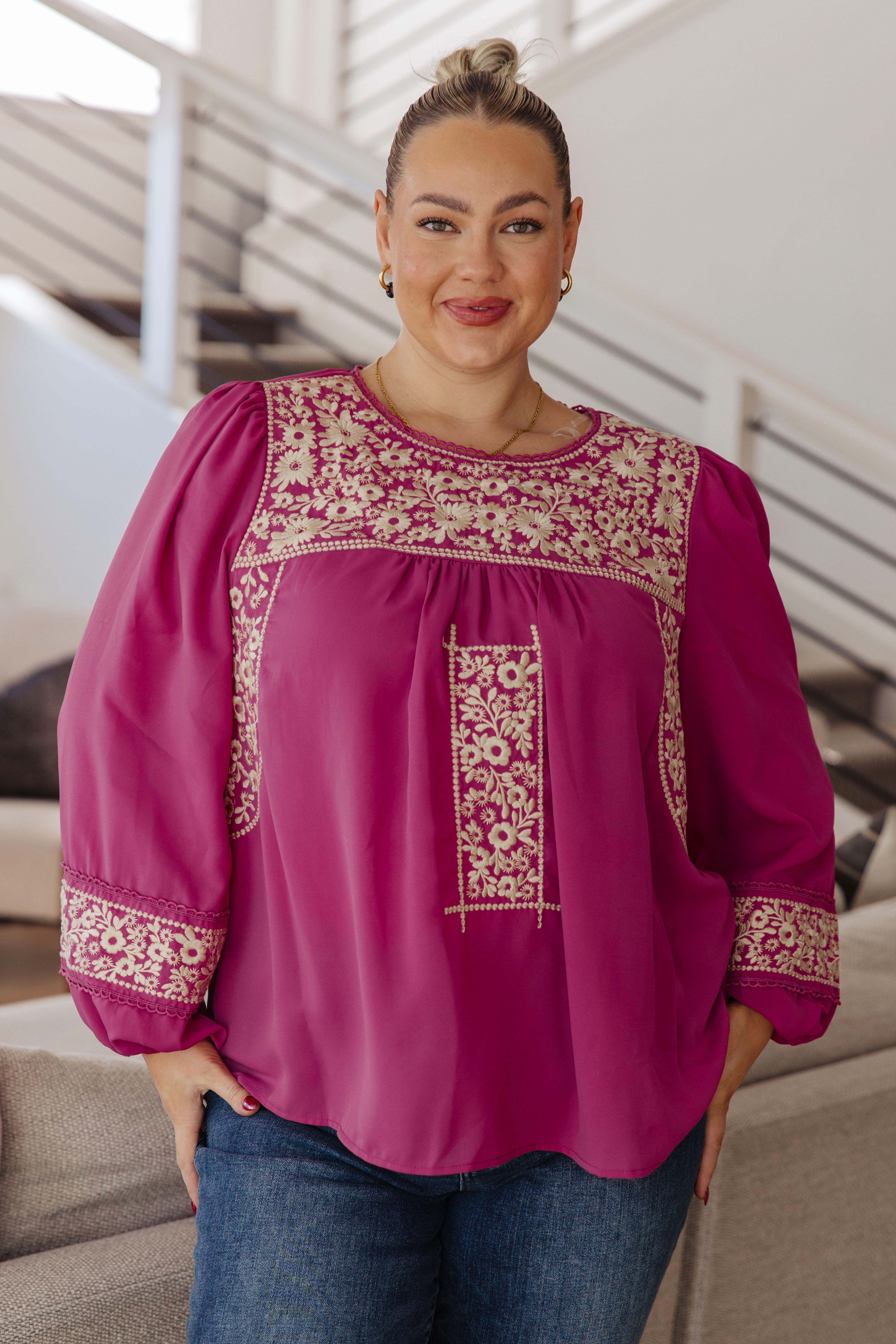Woman wearing a magenta long-sleeve blouse with intricate white embroidery and blue jeans standing in an indoor setting.