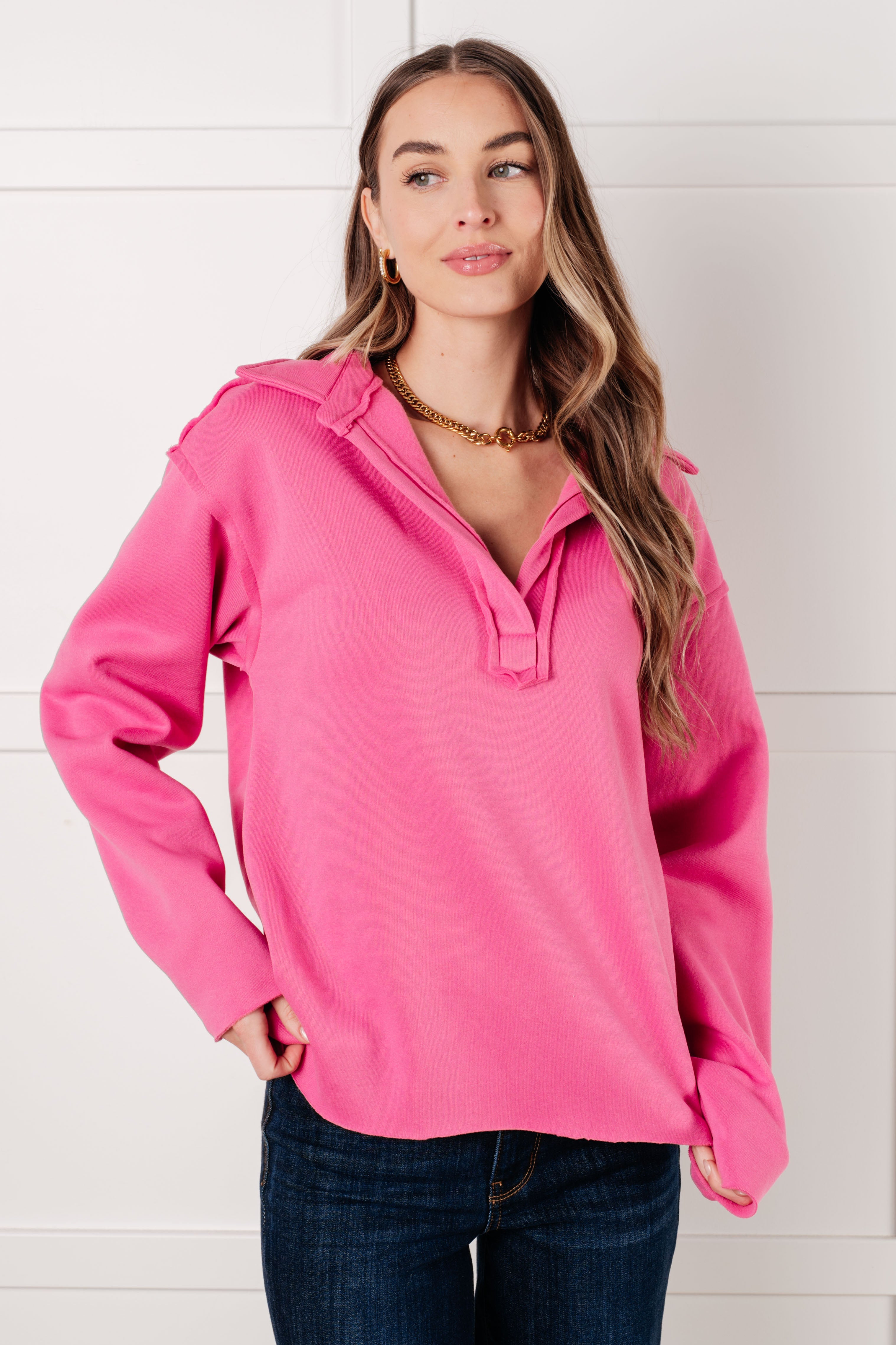 Woman wearing a hot pink colored pullover with exposed seams and dark blue jeans with a white background.