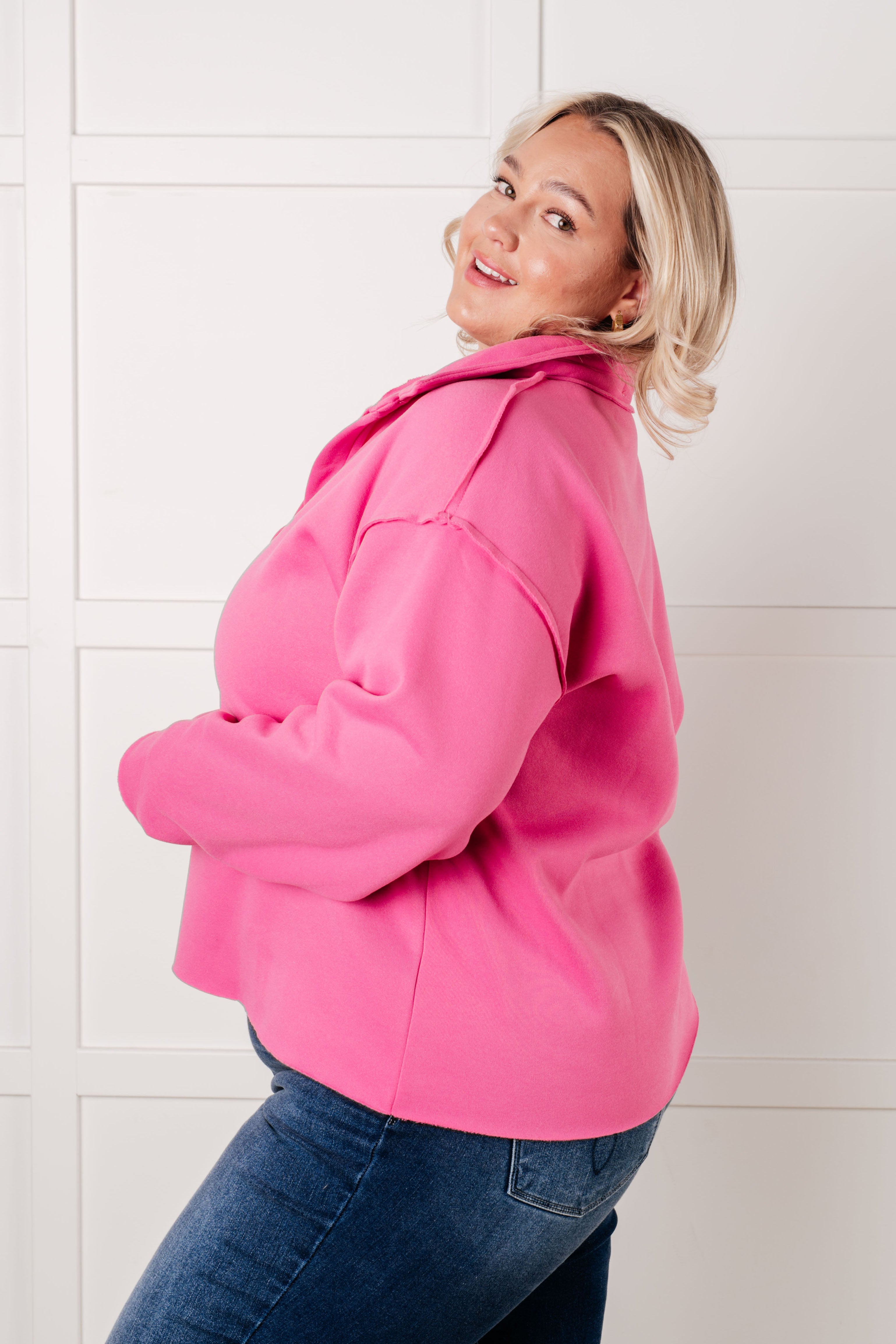 Woman wearing a hot pink colored pullover with exposed seams and blue jeans with a white background.