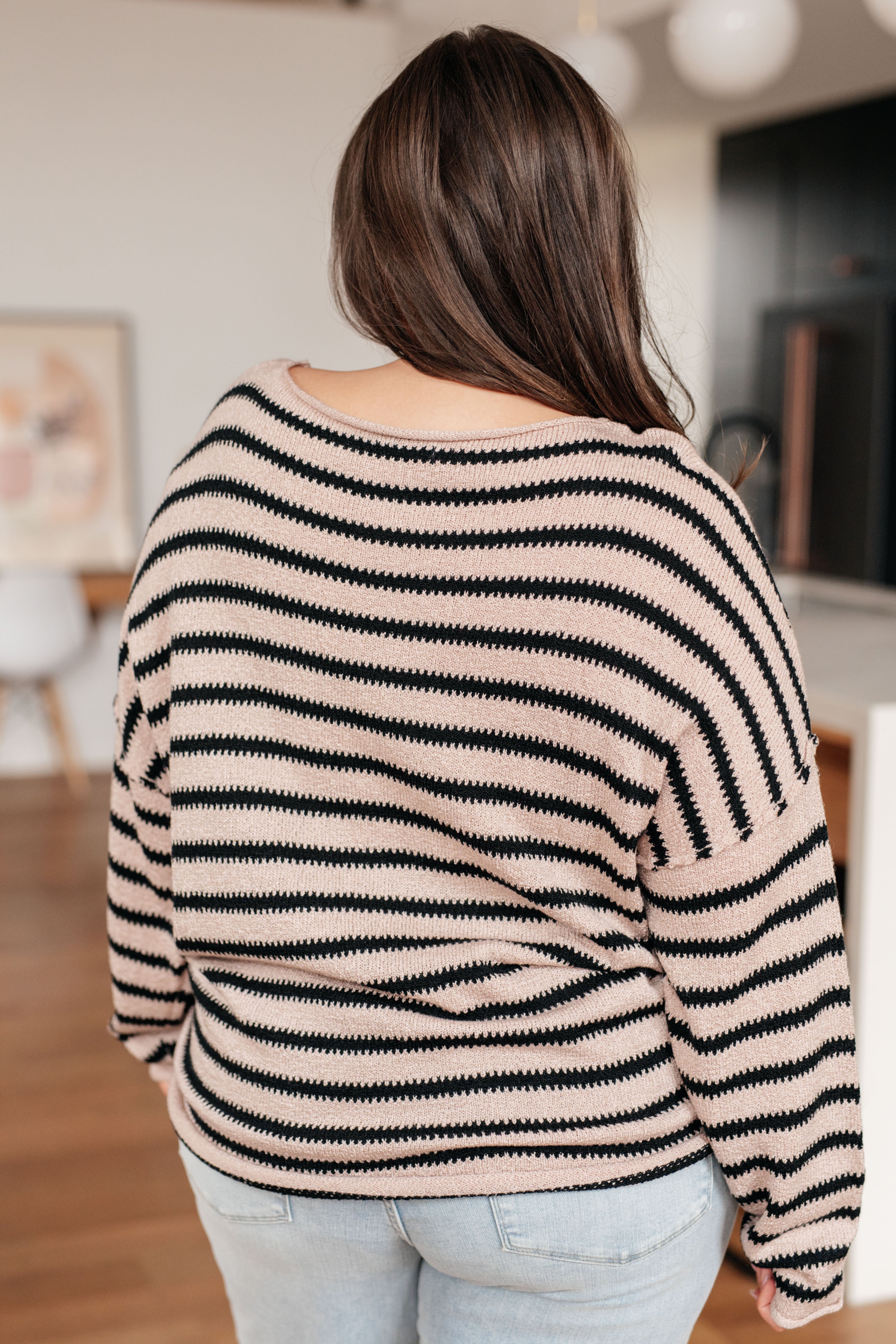 Woman wearing a taupe sweater with black stripes and light blue jeans indoors.