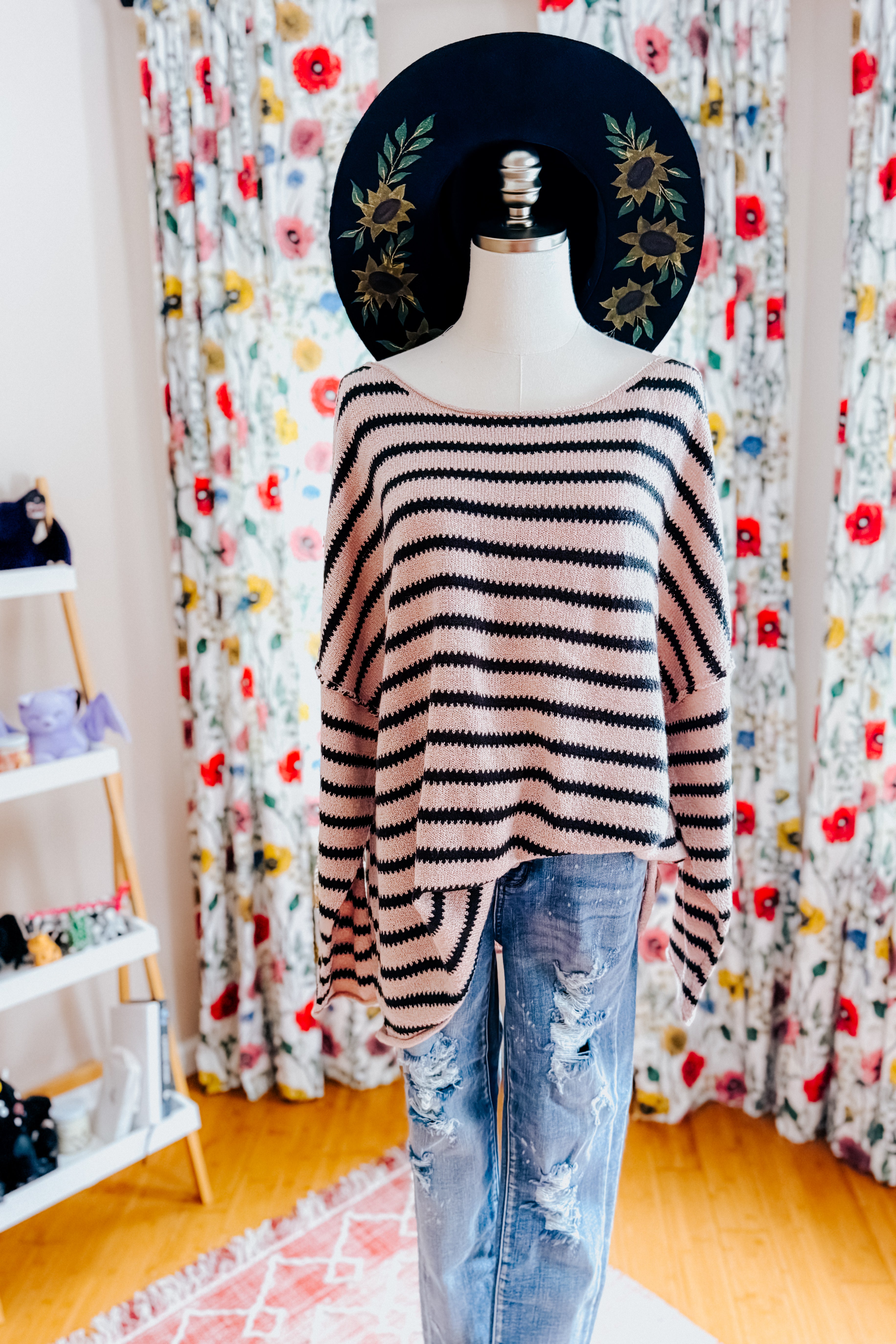A dress form wearing a taupe long sleeve sweater with black stripes, blue jeans and a painted wide brim hat in an indoor setting.