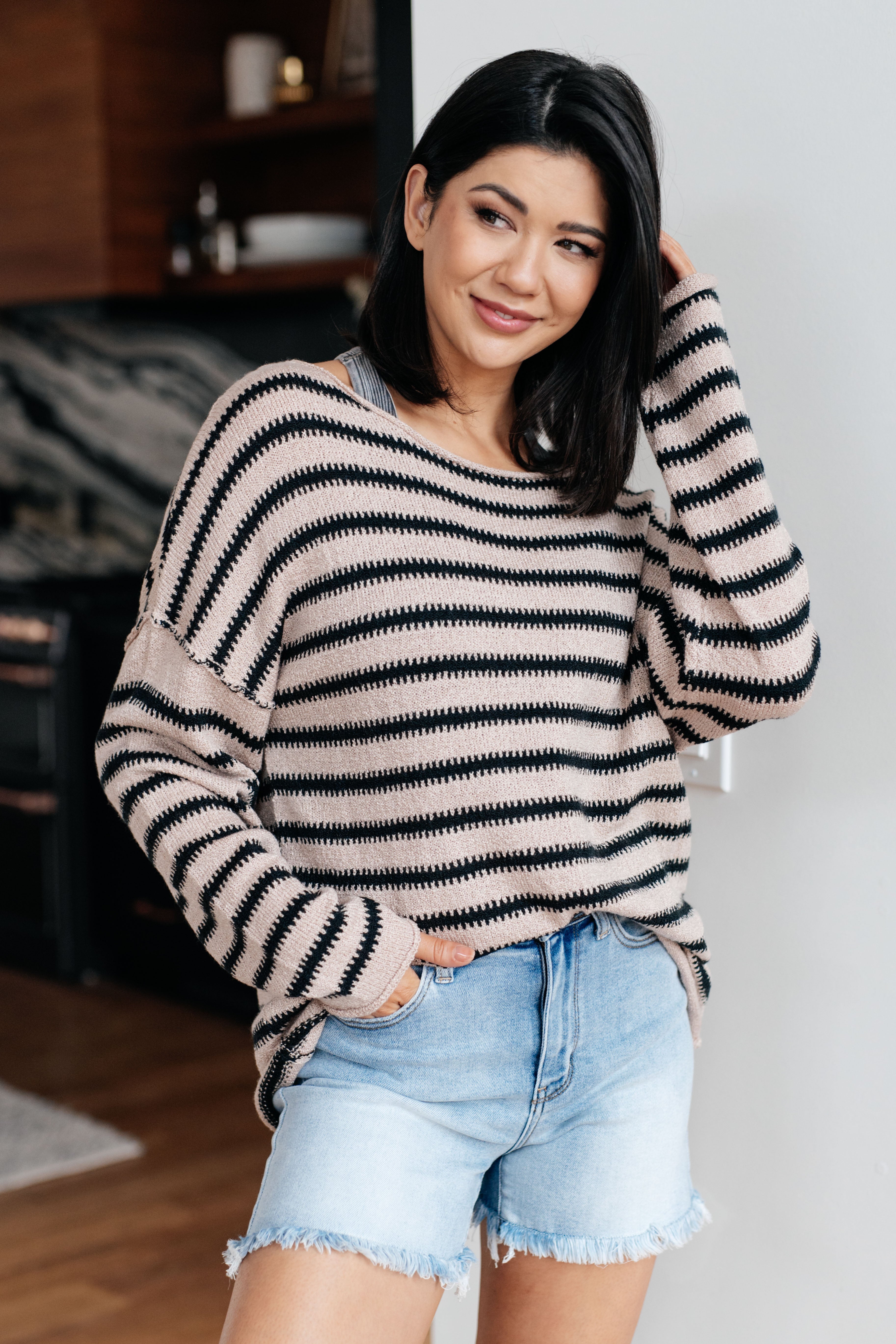 Woman wearing a taupe sweater with black stripes and denim shorts indoors.