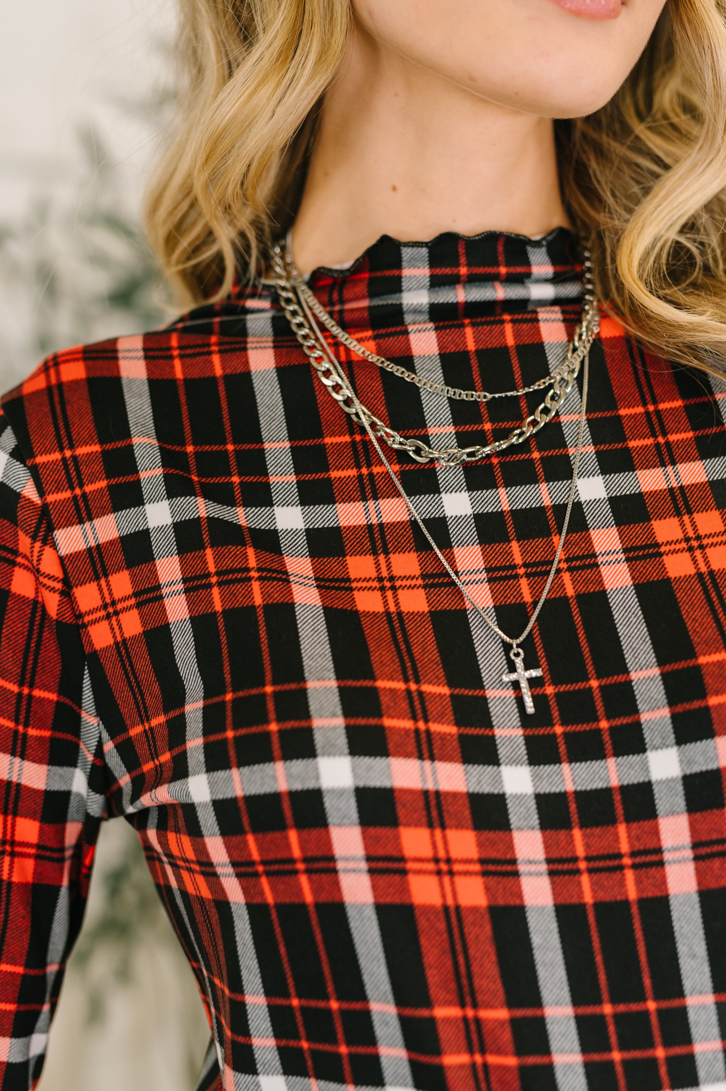 Close-up of a woman wearing a red and black plaid sheer shirt with layered necklaces, including one with a cross pendant.
