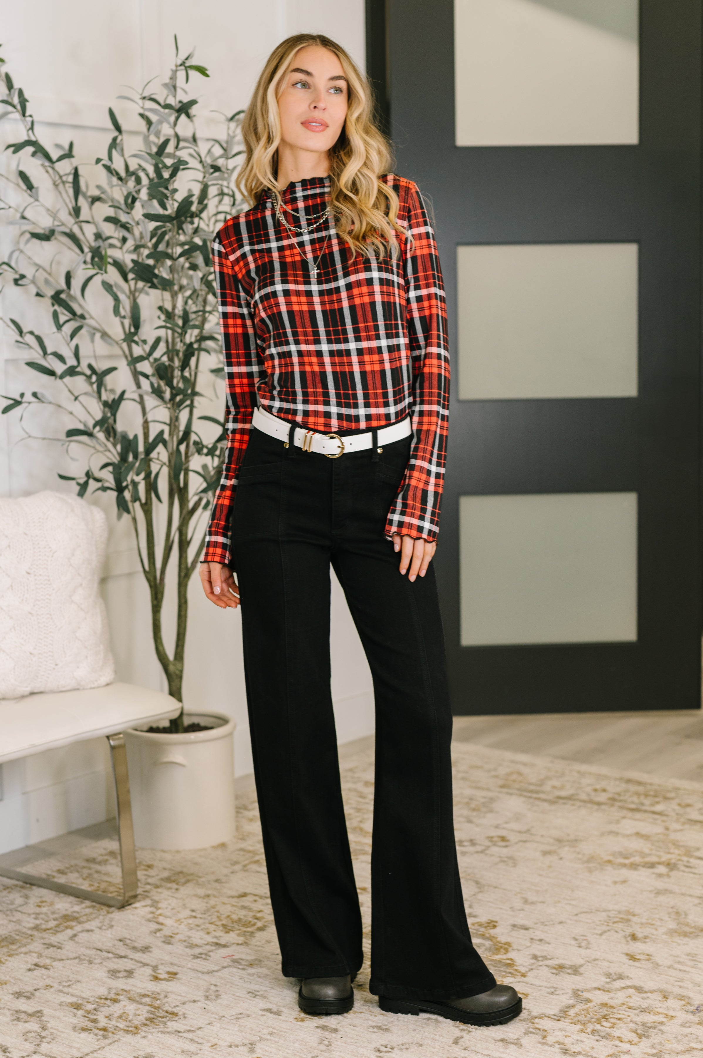 Woman wearing a red and black sheer plaid shirt with black pants and a white belt and black boots standing in an indoor setting.