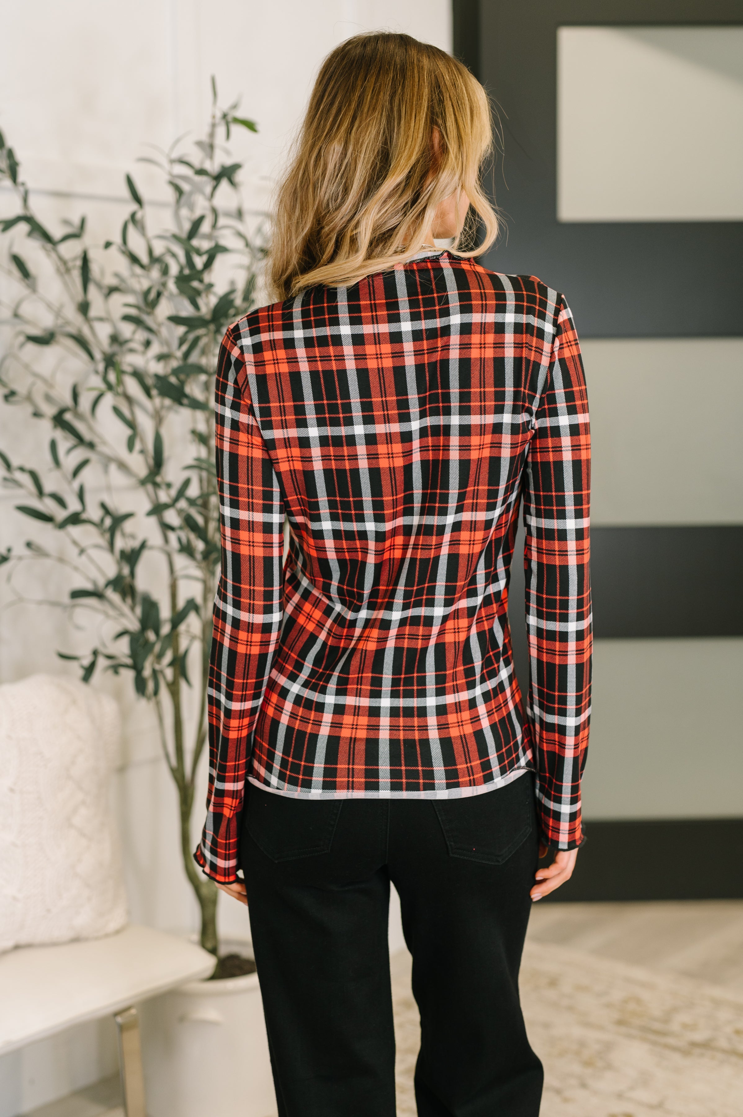 Woman with her back facing the camera wearing a red and black sheer plaid shirt over back jeans in an indoor setting