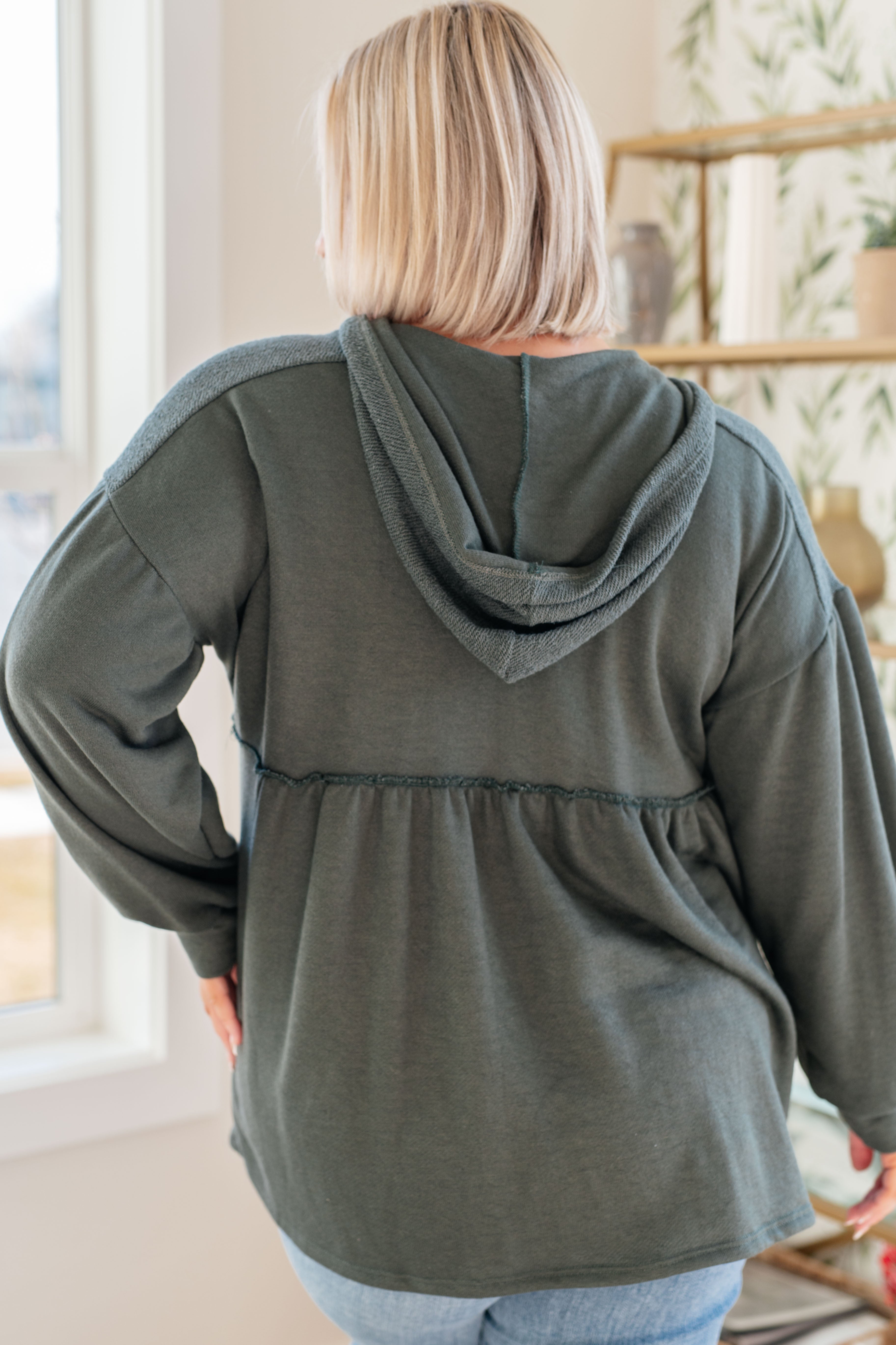 Woman wearing a dusty green v-neck hooded pullover and blue jeans in an indoor setting.