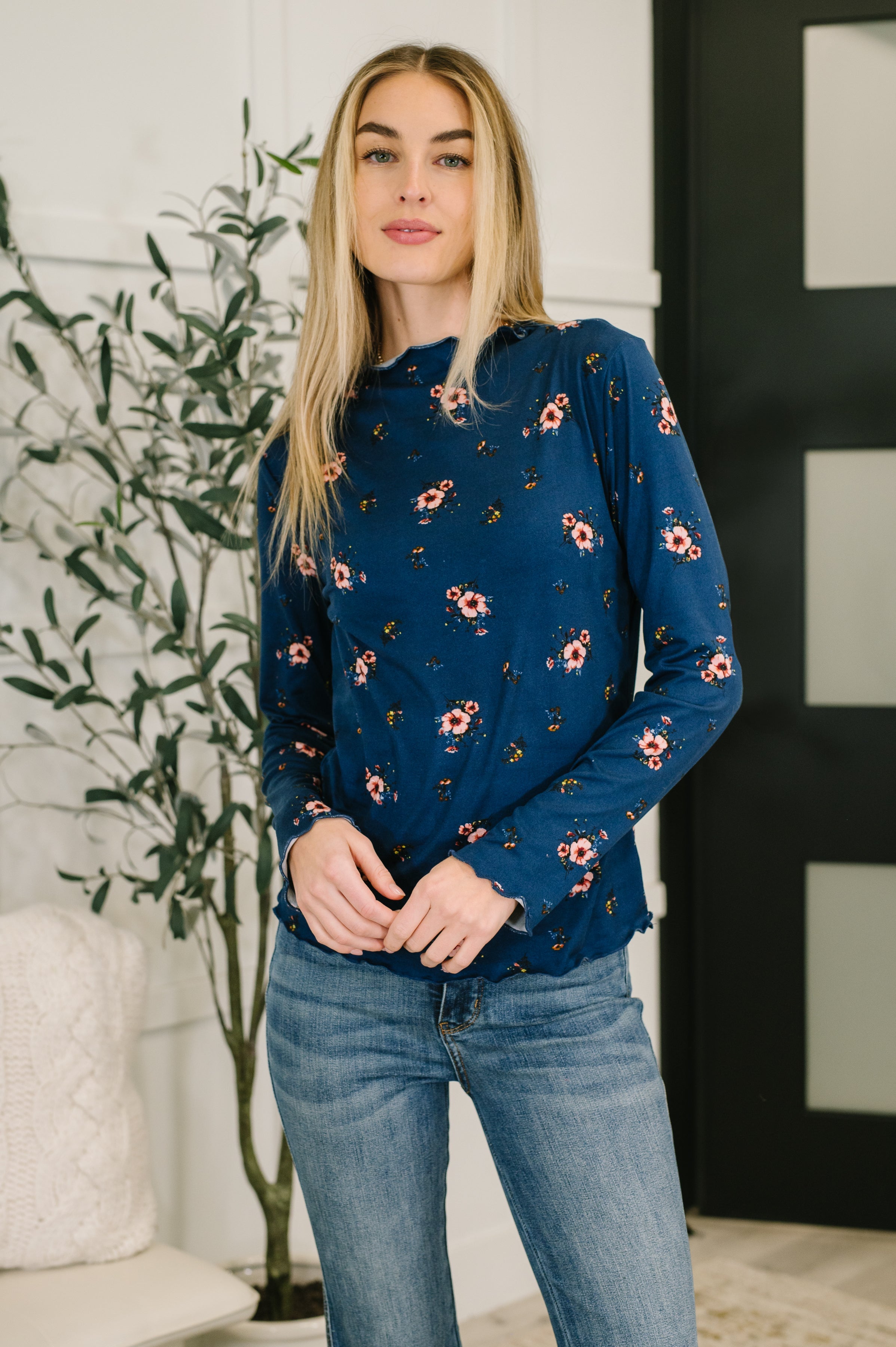 Woman wearing a blue floral top and blue jeans standing indoors with a plant in the background