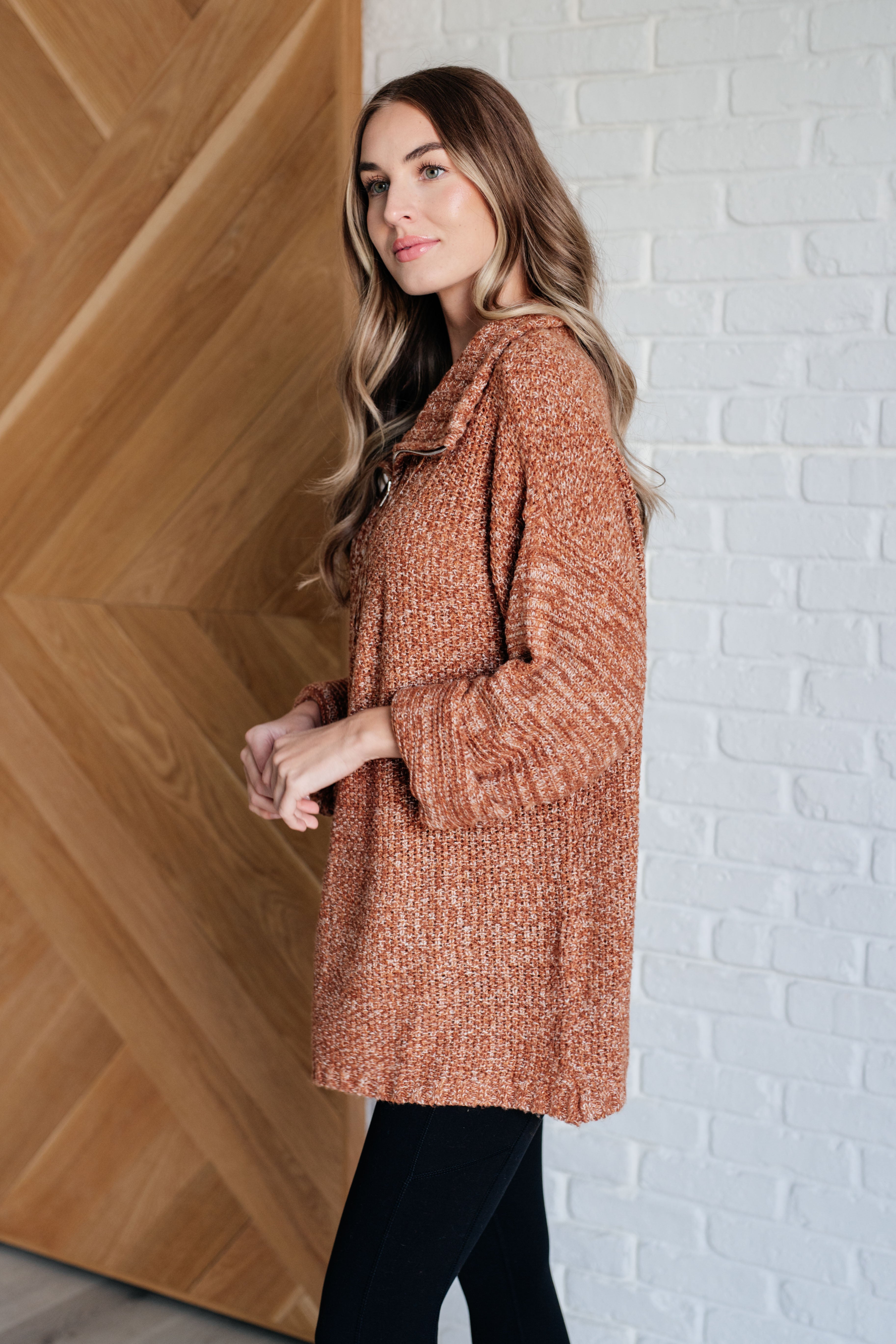 Woman wearing a rust colored oversized long-sleeve sweater and black leggings standing against a white brick wall.
