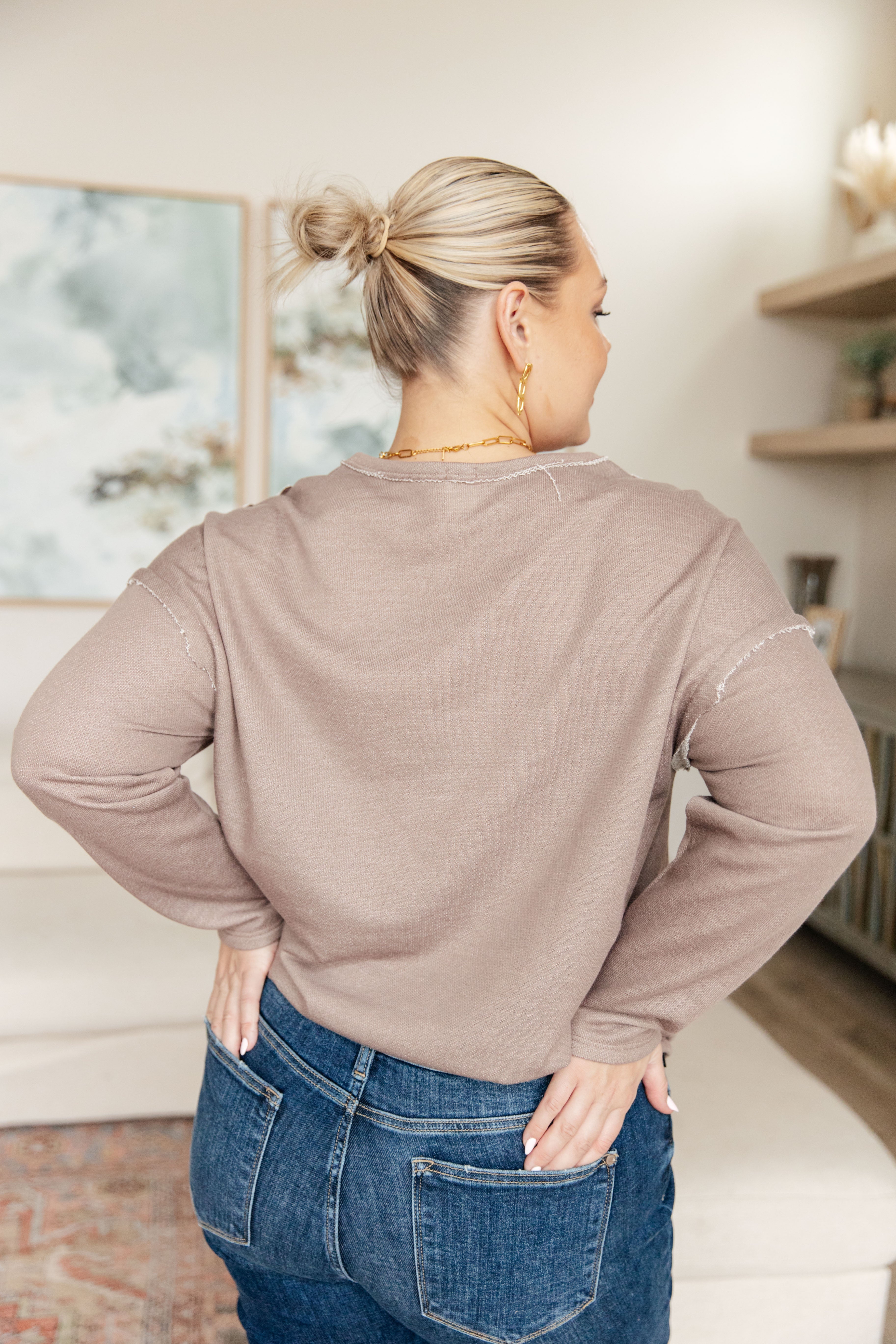 Woman wearing a taupe reverse stitch v-neck pullover with raw accents and blue jeans standing in an indoor setting.