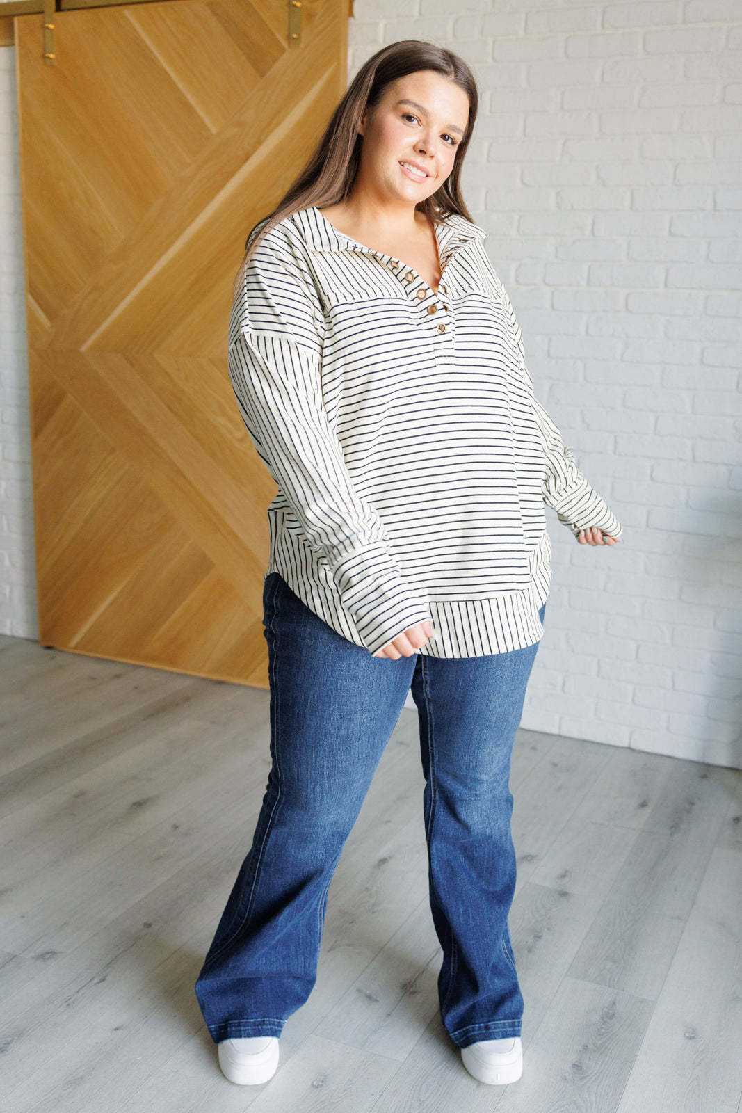 Woman wearing a white and black striped collared pullover top, blue jeans and white sneakers standing against a white brick wall.