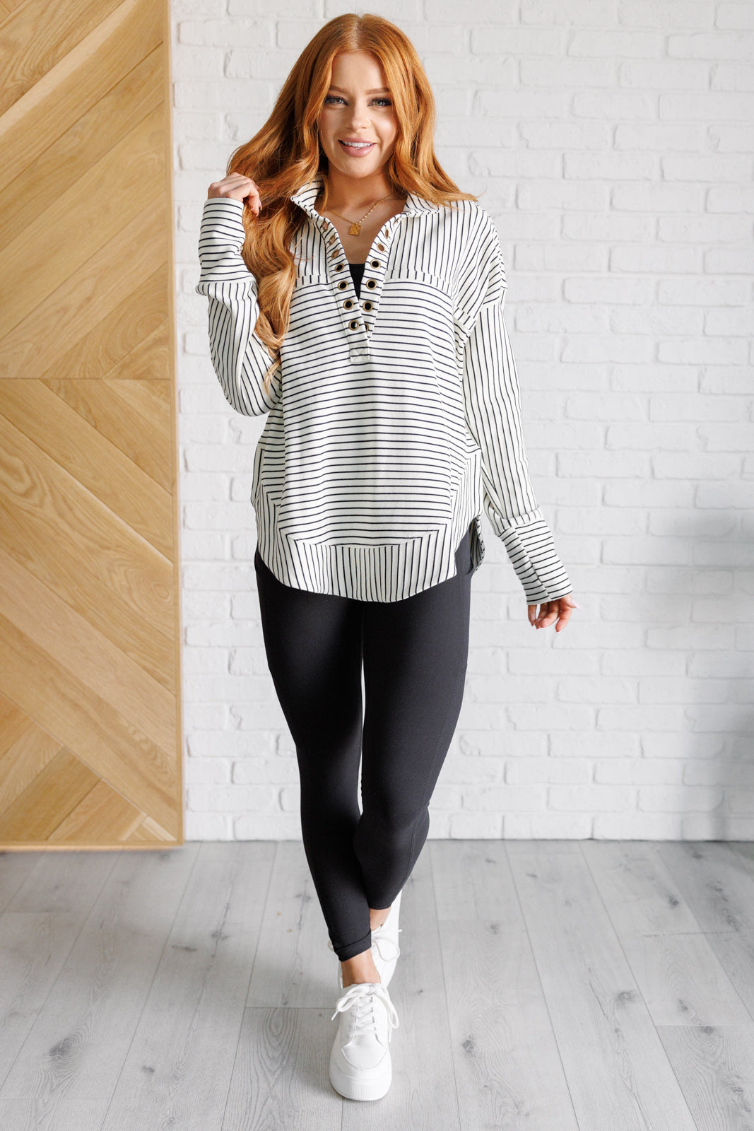 Woman wearing a white and black striped collared pullover top, black leggings and white sneakers standing against a white brick wall.