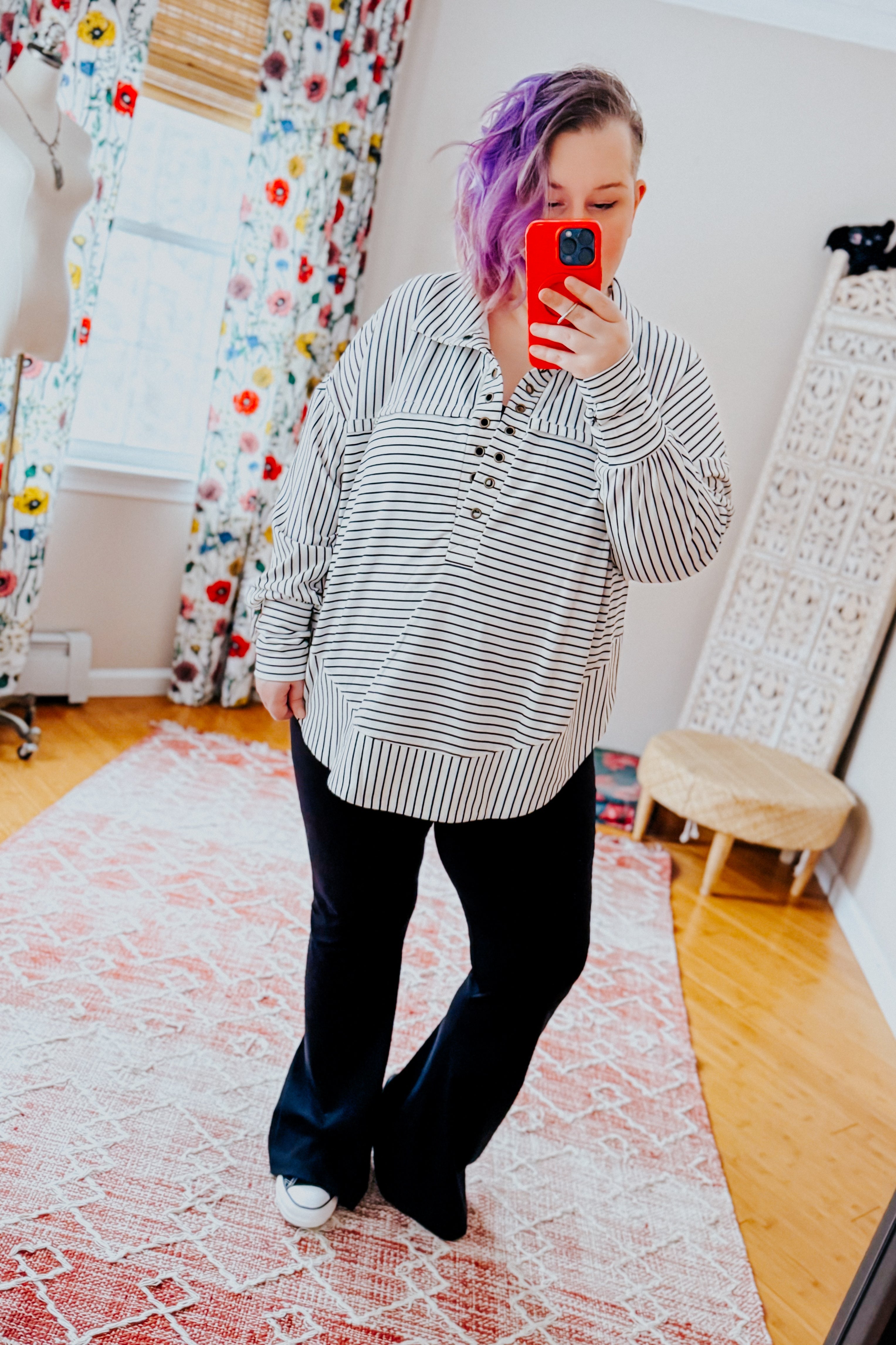 A woman taking a mirror selfie wearing a white and black striped collared pullover top, black pants and black converse sneakers standing in an indoor setting.