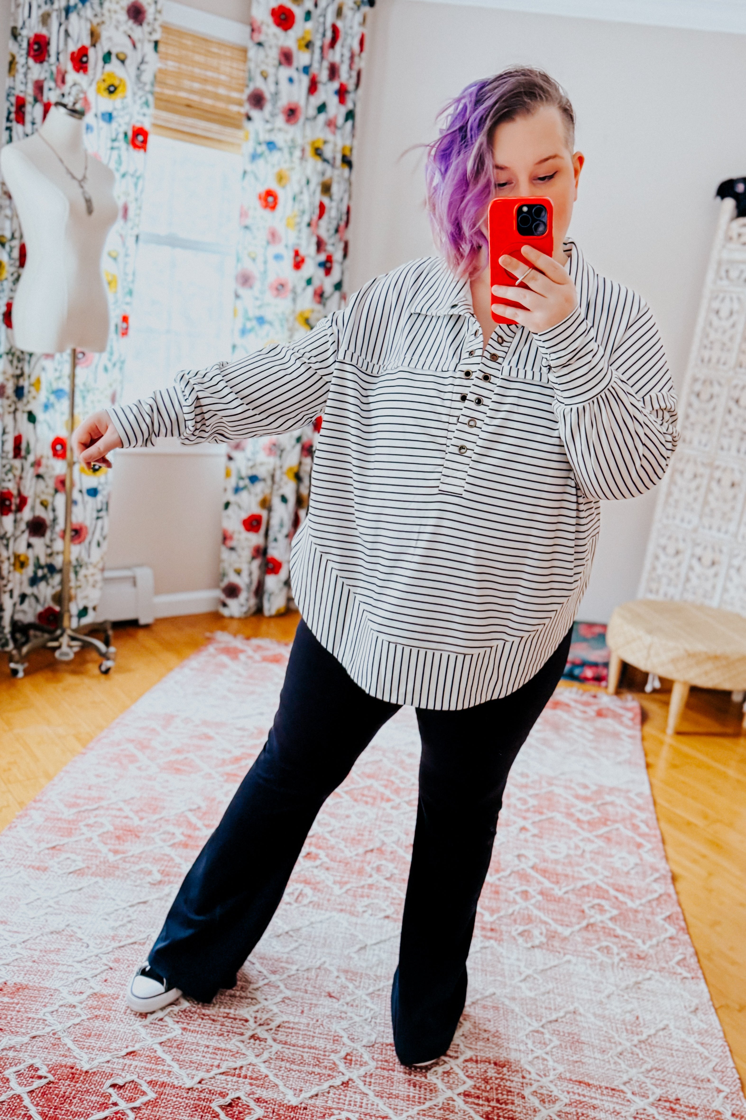 A woman taking a mirror selfie wearing a white and black striped collared pullover top, black pants and black converse sneakers standing in an indoor setting.
