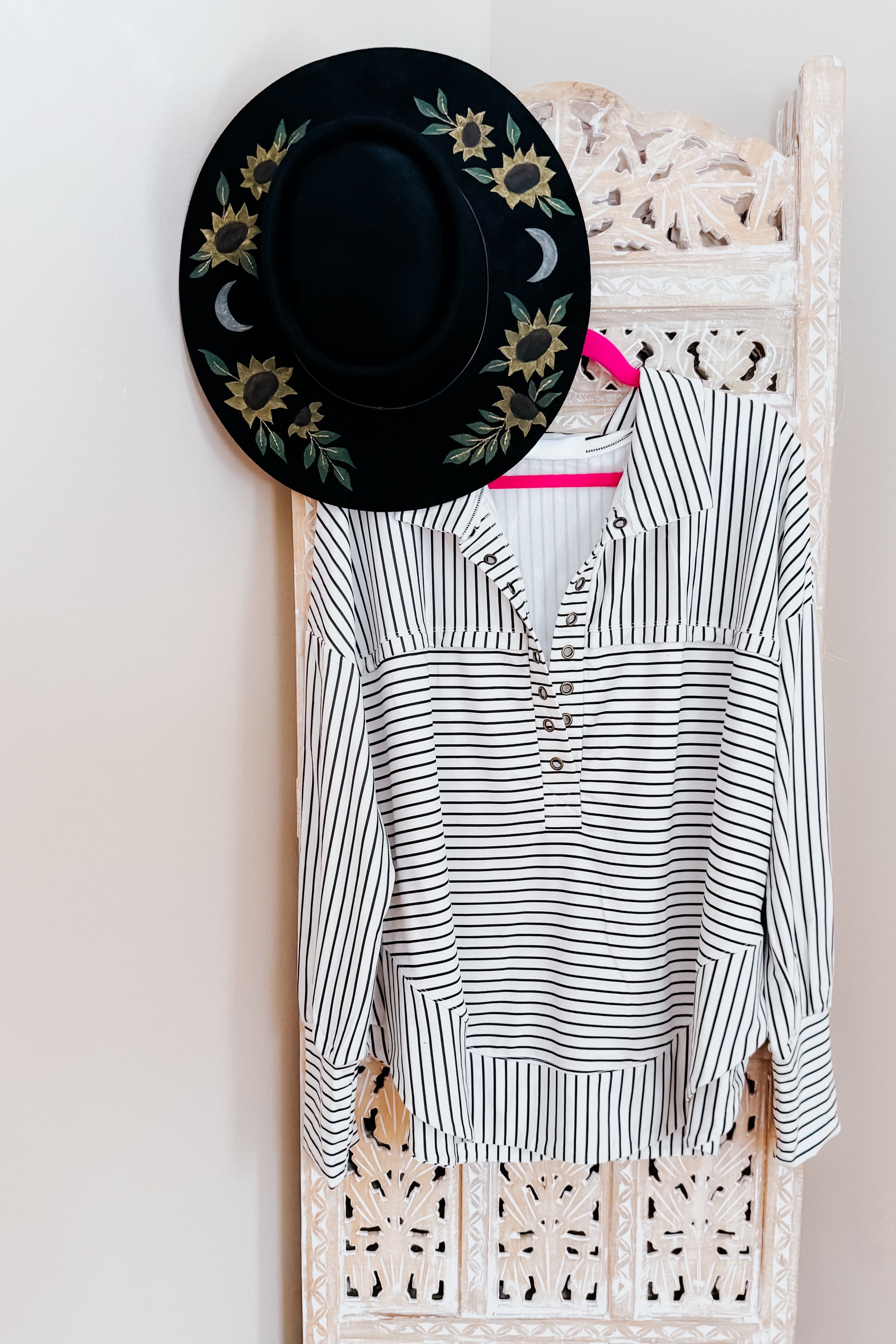 A white and black striped collared pullover top hanging from a room divider from a bright pink hanger with a black hand painted black large brim hat.