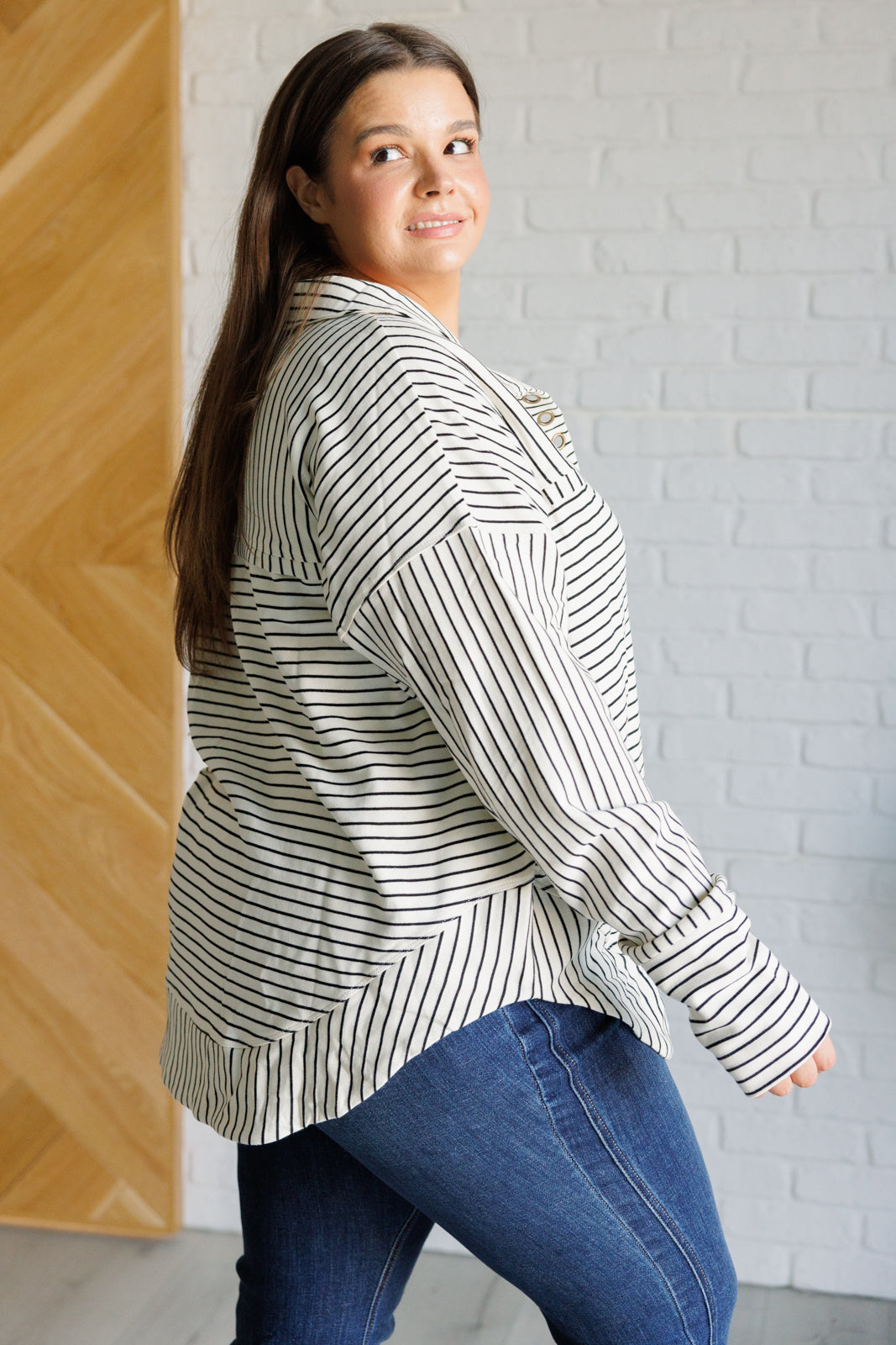 Woman wearing a white and black striped collared pullover top and blue jeans standing against a white brick wall.