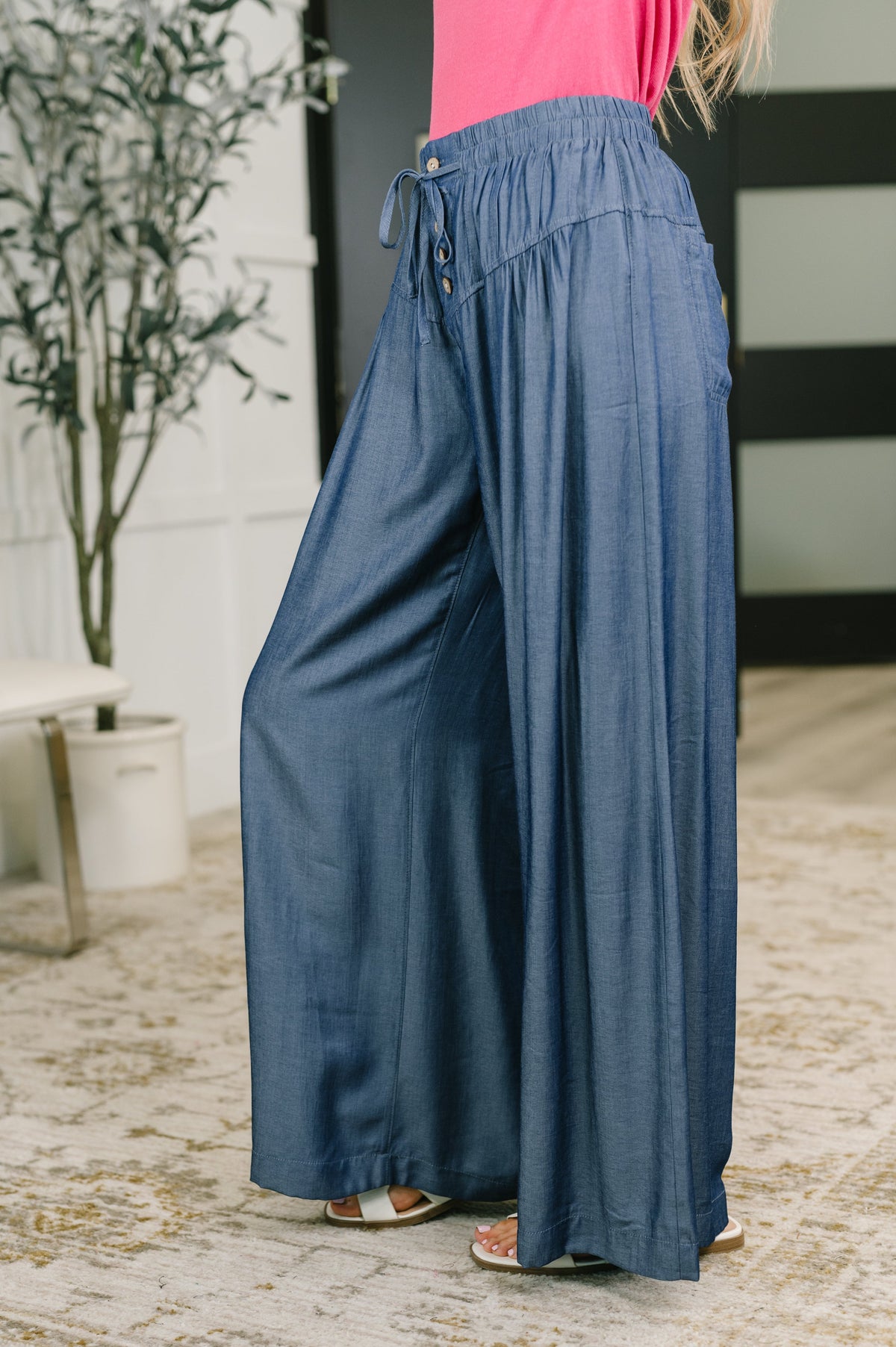Woman wearing blue high waisted wide leg bottoms with a gathered waistband over a pink top indoors