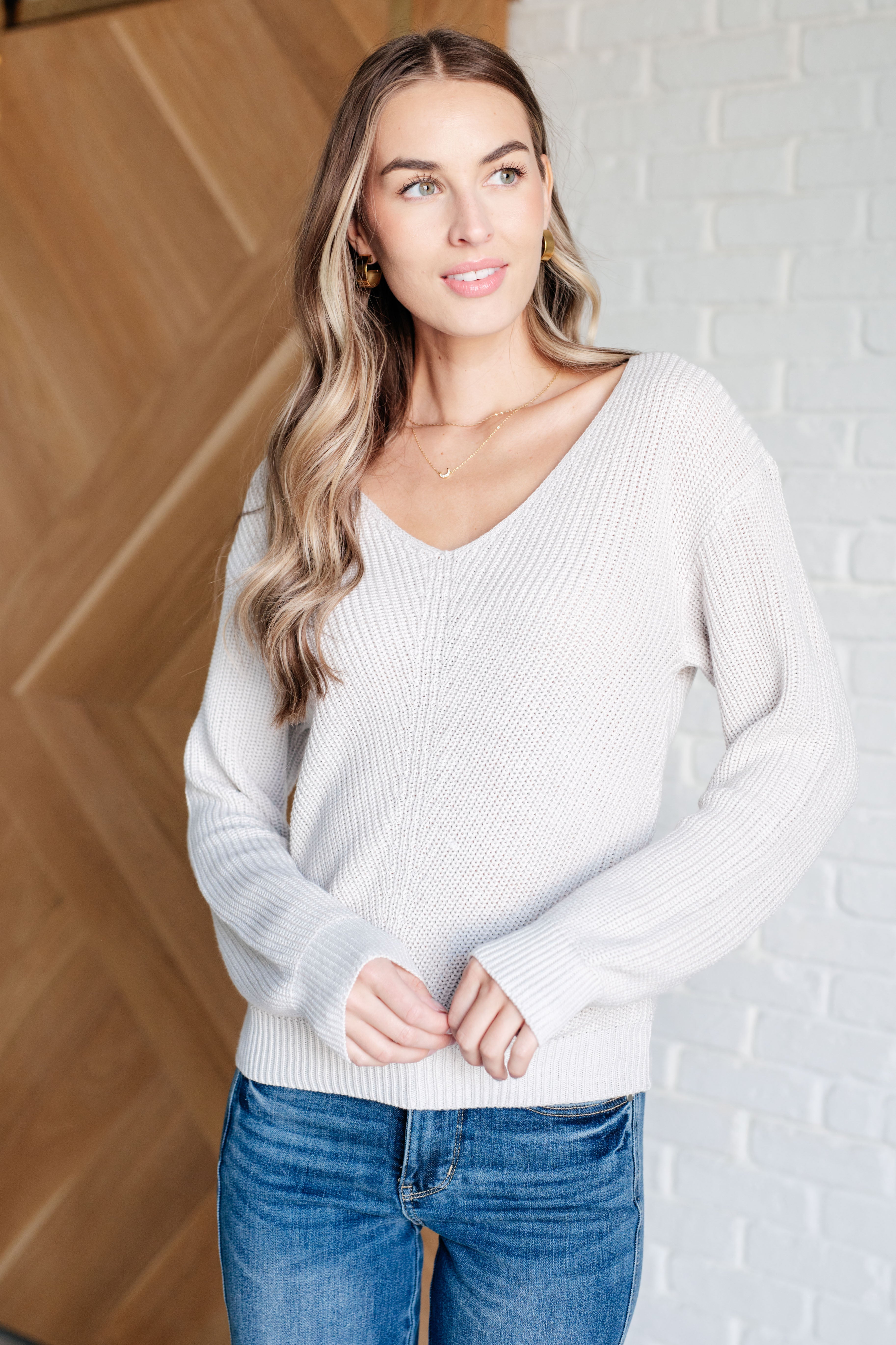 Woman wearing a light grey knit v-neck sweater and blue jeans standing against a white brick wall.