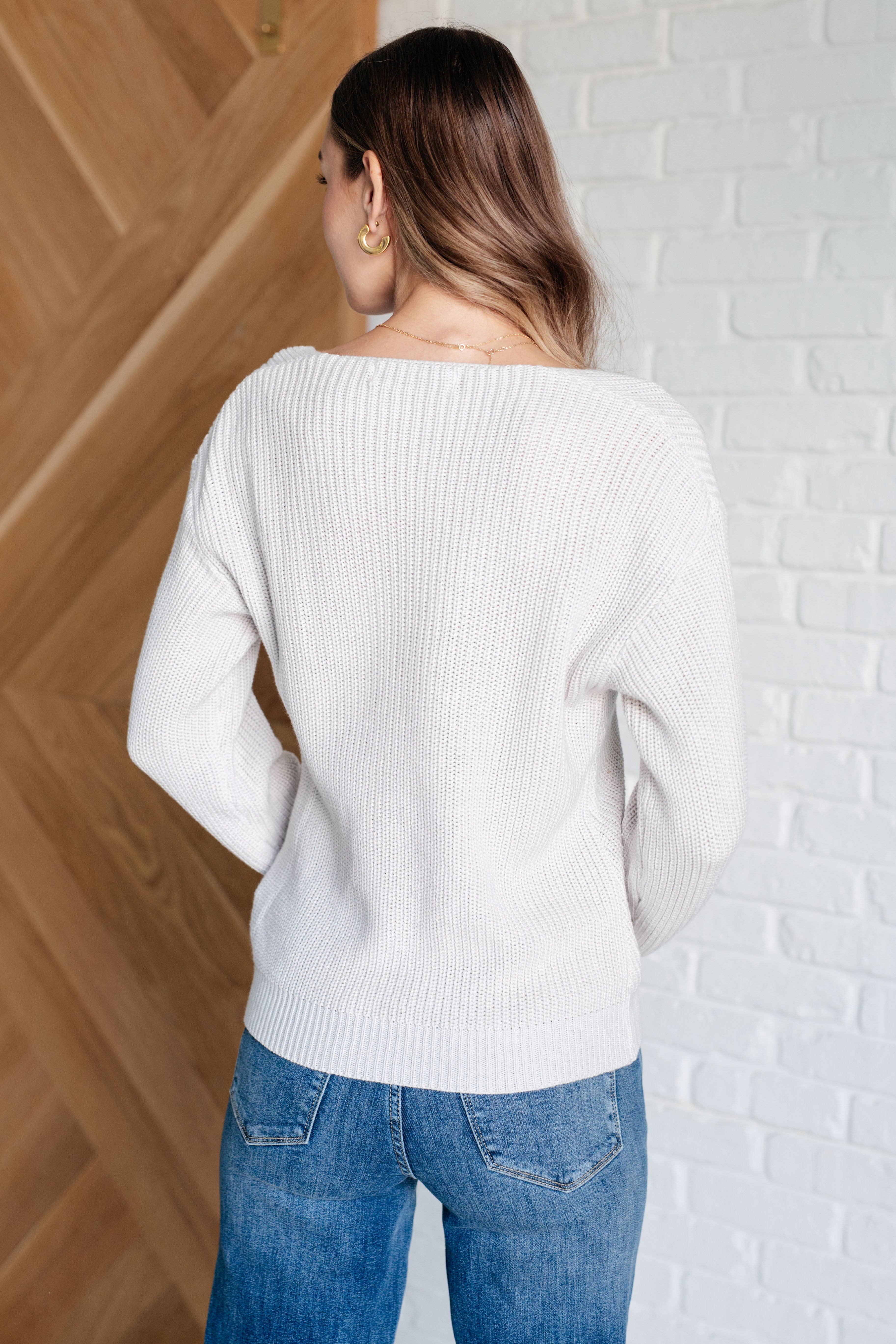 Woman wearing a light grey knit v-neck sweater and blue jeans standing against a white brick wall.