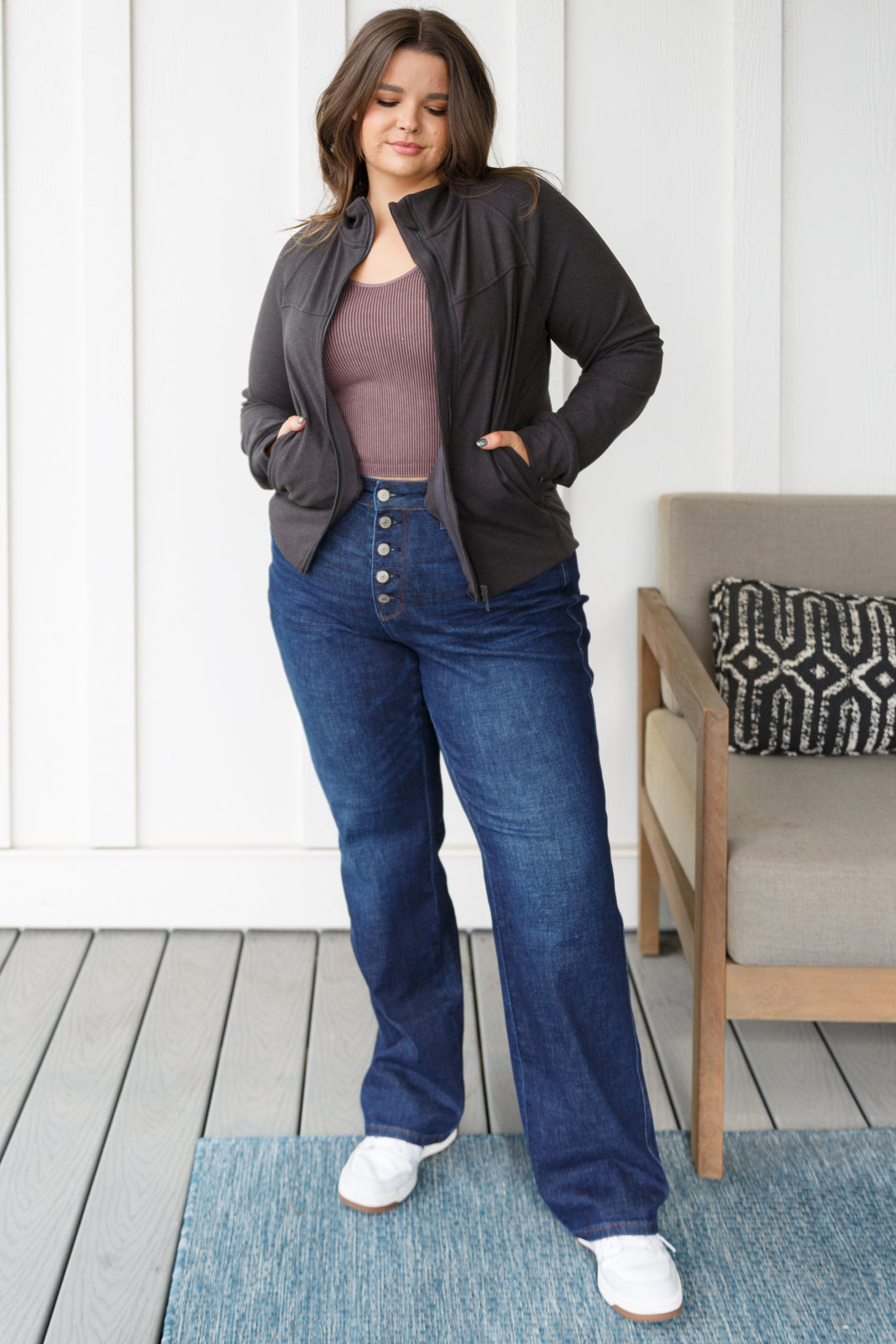 Woman wearing a dark gray jacket, brown top, and blue jeans on a wooden deck.