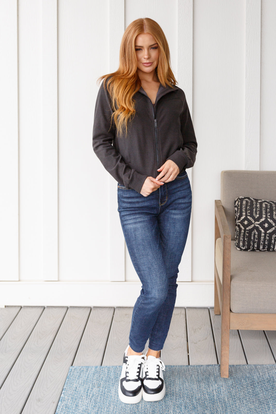 Woman wearing a gray jacket, blue jeans, and sneakers standing on a wooden deck.