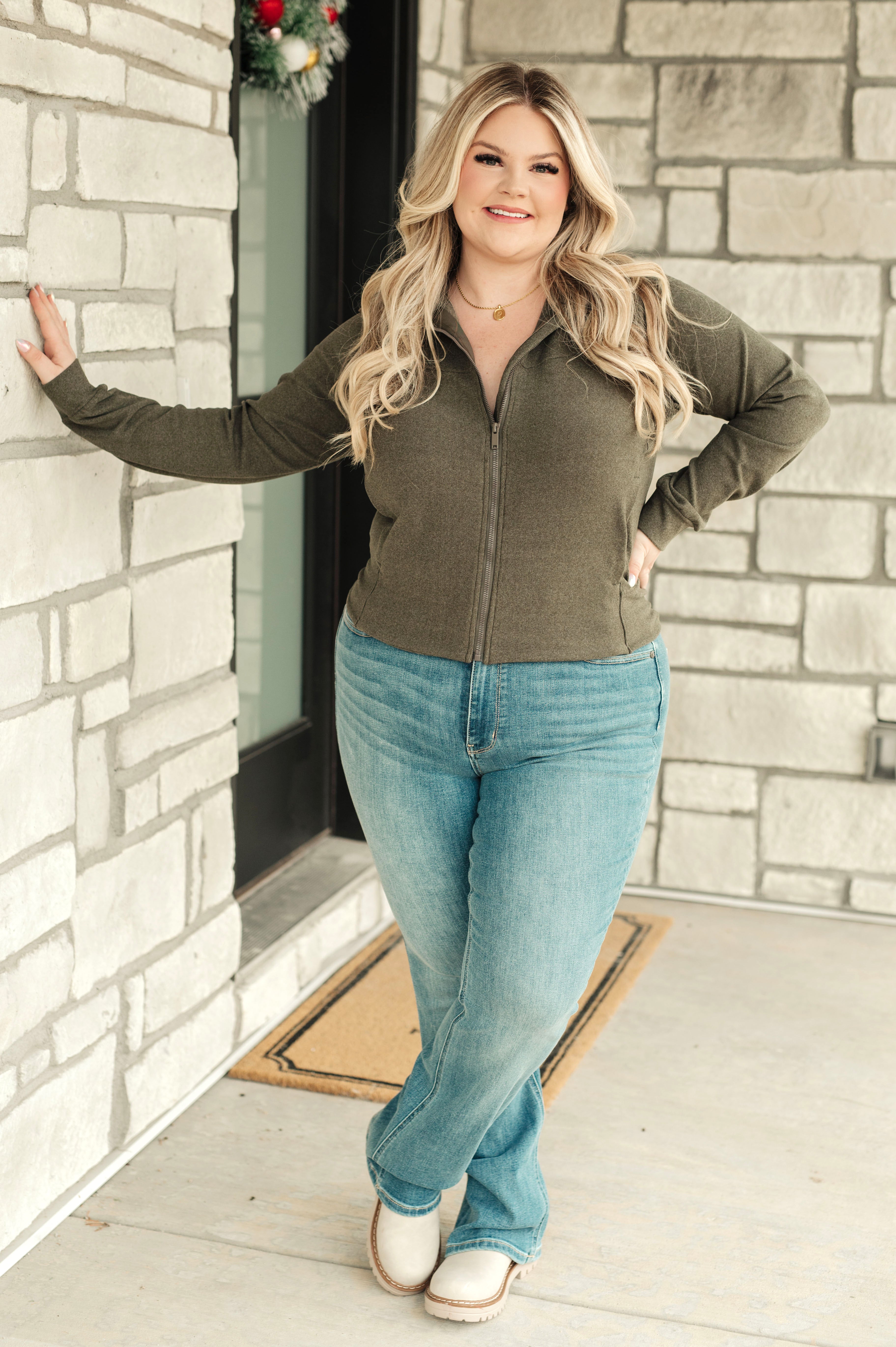 Woman wearing an olive zip-up jacket, blue jeans and beige boots standing against a stone wall.