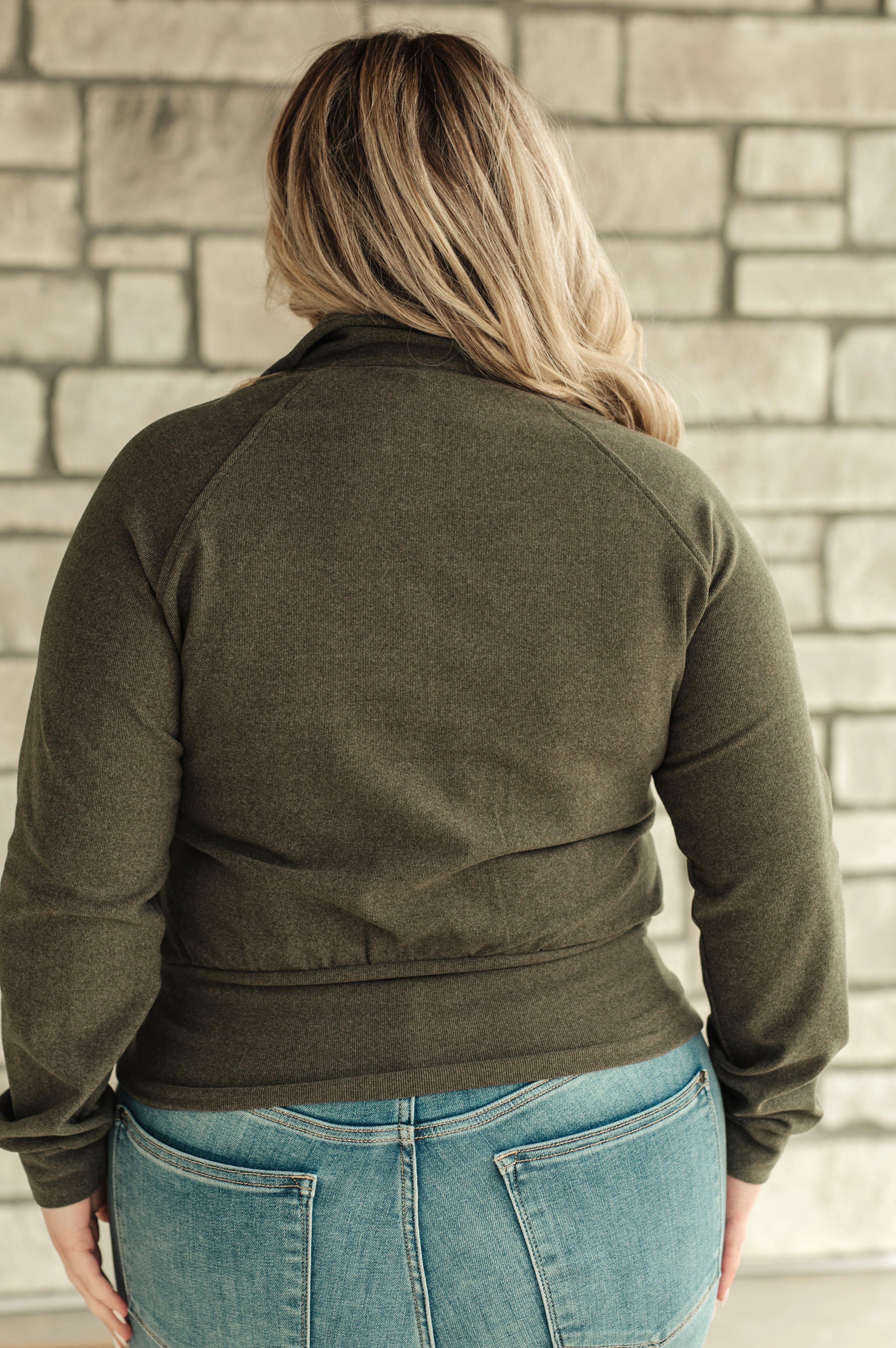 Woman wearing an olive green zip-up jacket and blue jeans against a stone wall.