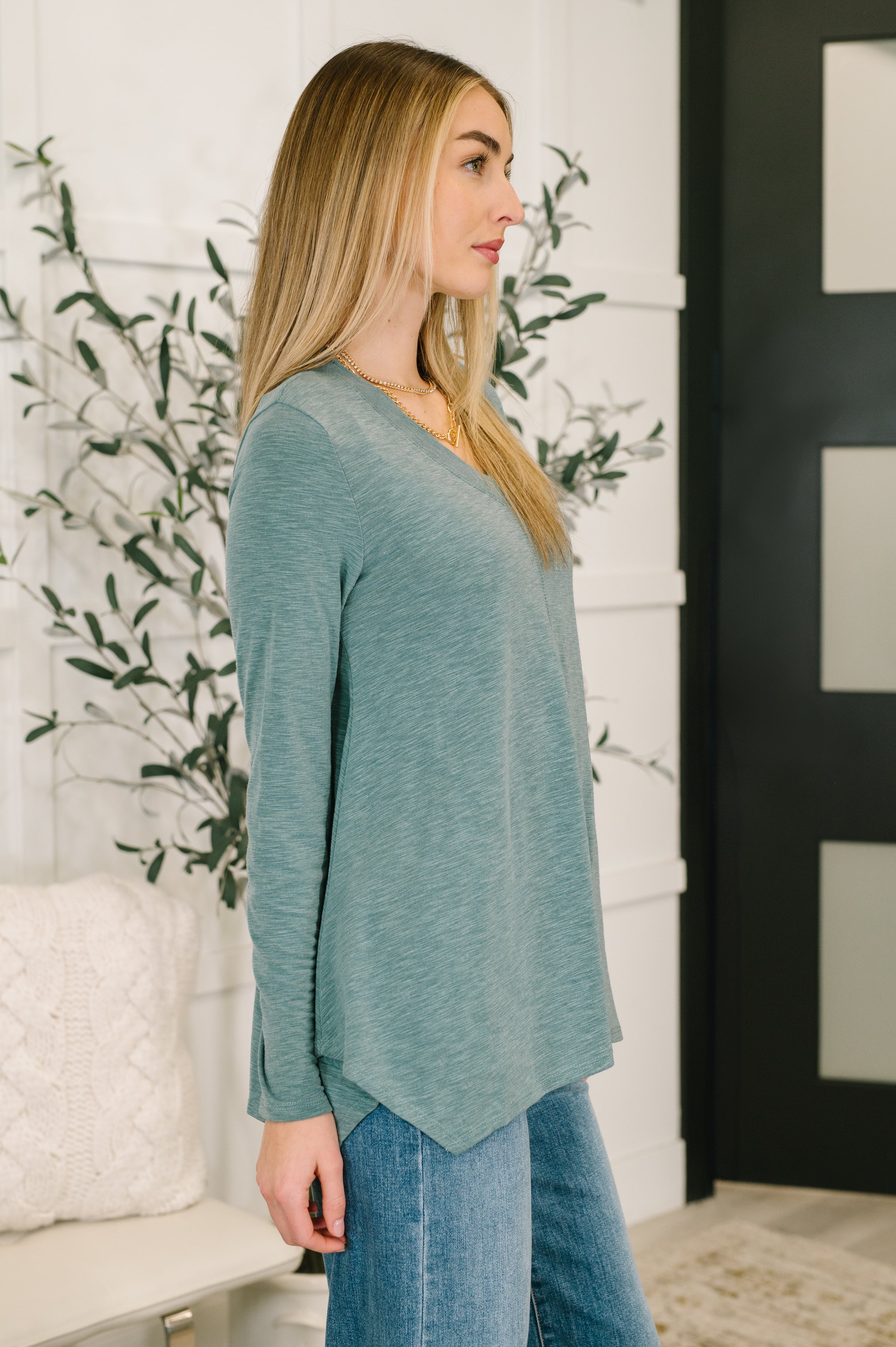 Woman wearing a light teal v neck long sleeve top and blue jeans standing in an indoor setting.