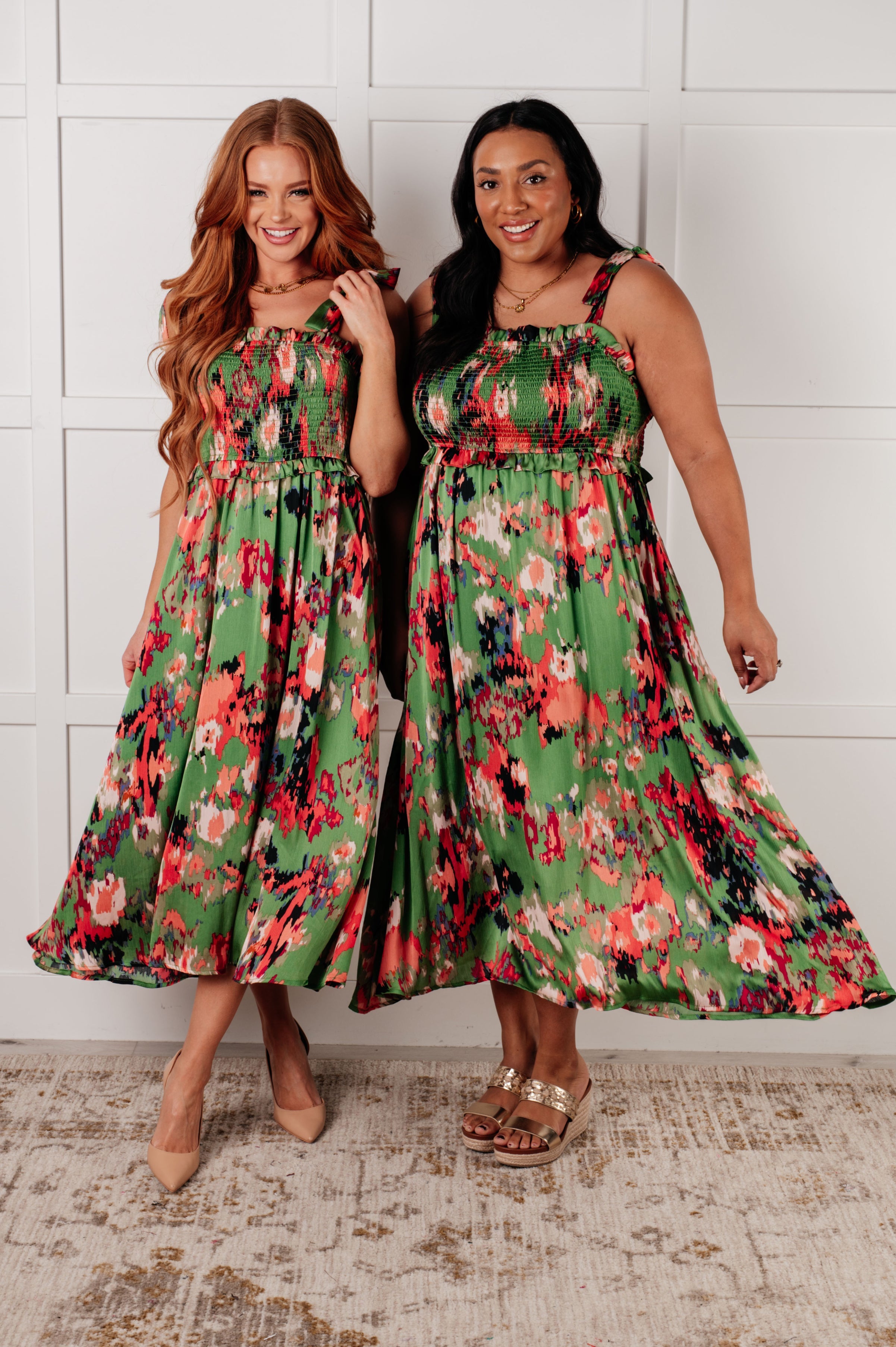 Two women wearing matching green floral smocked midi dresses with wide shoulder straps standing side by side indoors