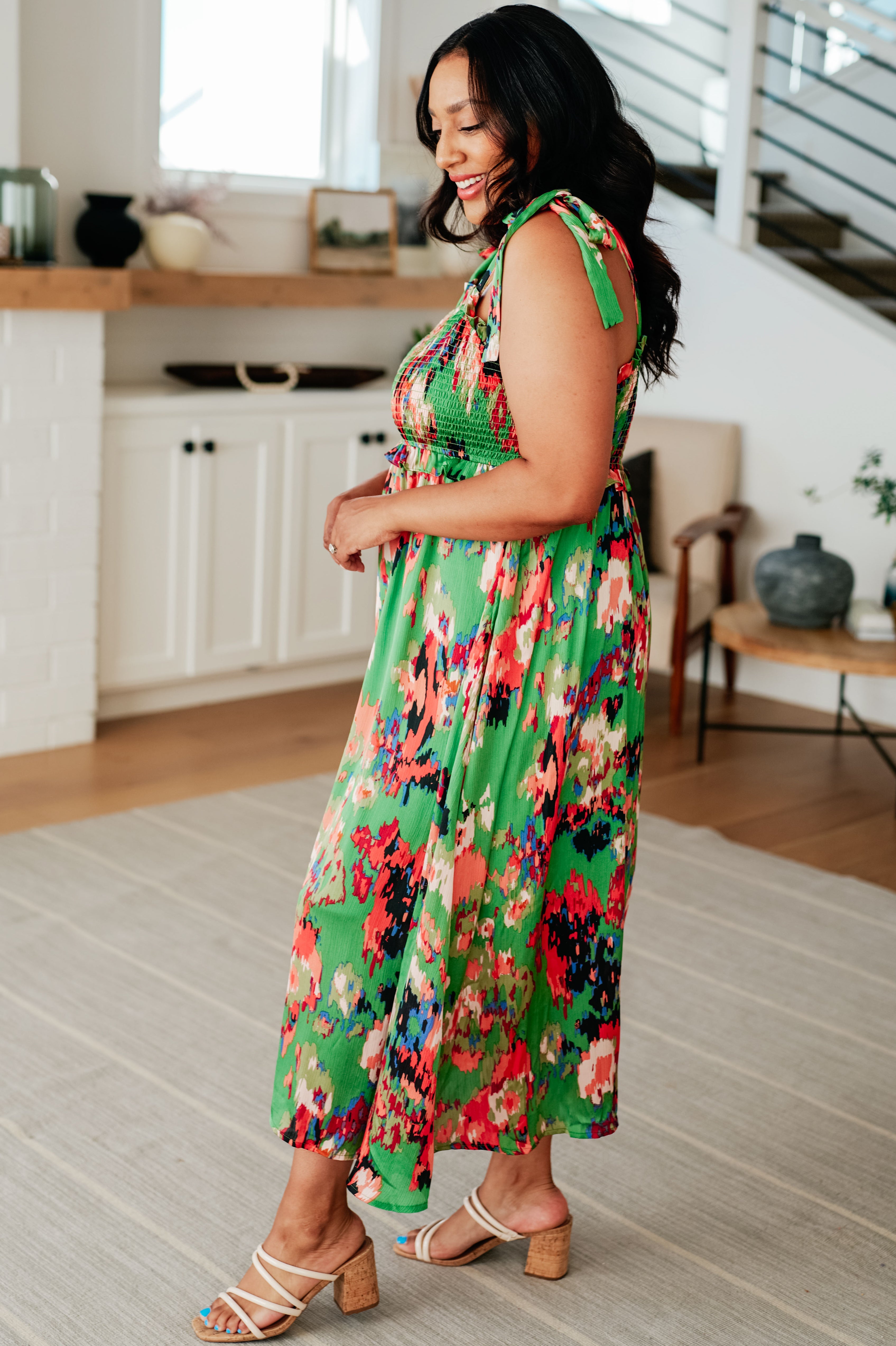 Woman in flowy green floral dress with smocked bodice and tied shoulder straps standing indoors side view