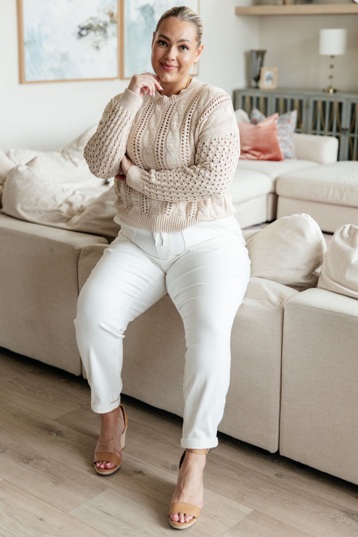 Woman wearing a cream cable knit sweater with openwork details and white pants while seated on a sofa in a bright living room