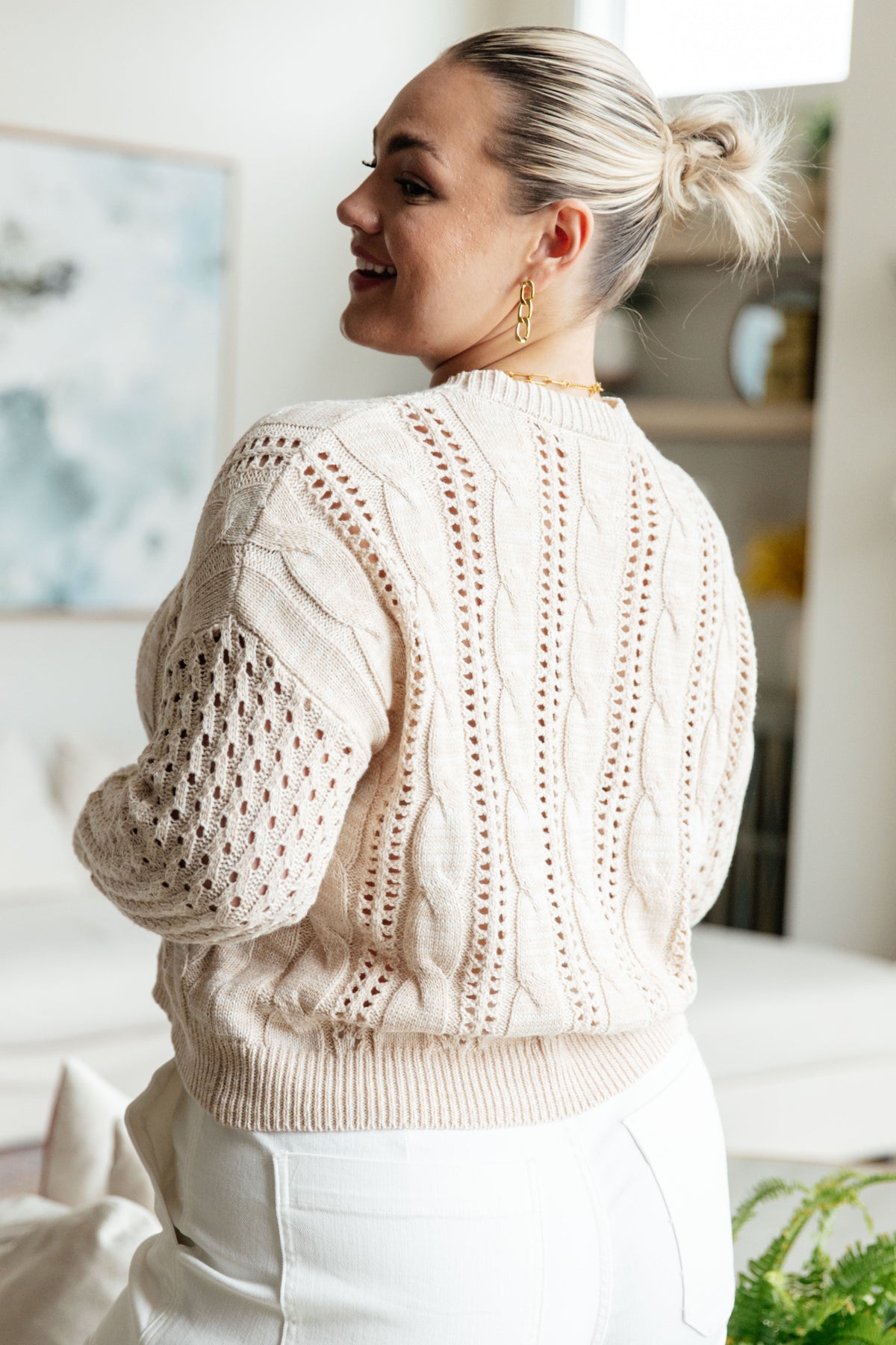 Back view of a woman wearing a cream cable knit sweater with openwork details and ribbed trim paired with white pants indoors