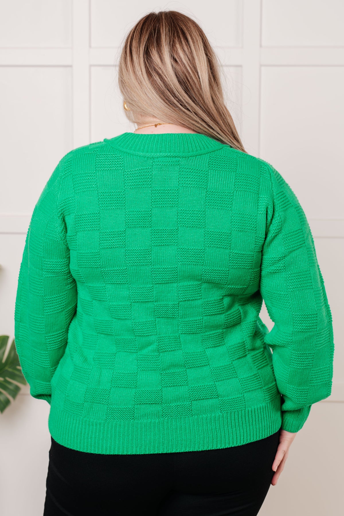 Back view of a woman wearing a green monochromatic checkered sweater knit pullover with ribbed trim and black pants indoors
