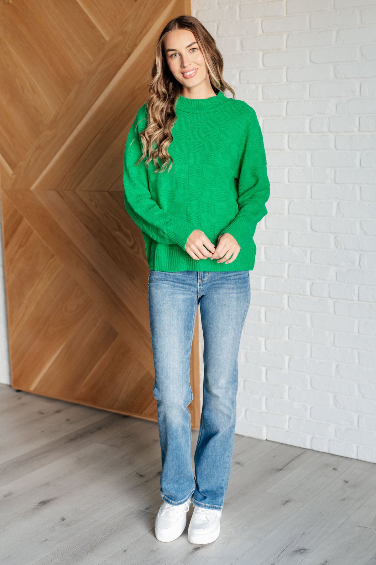 Woman modeling a green sweater knit pullover with ribbed trim, blue jeans, and white sneakers against a wood and brick wall backdrop