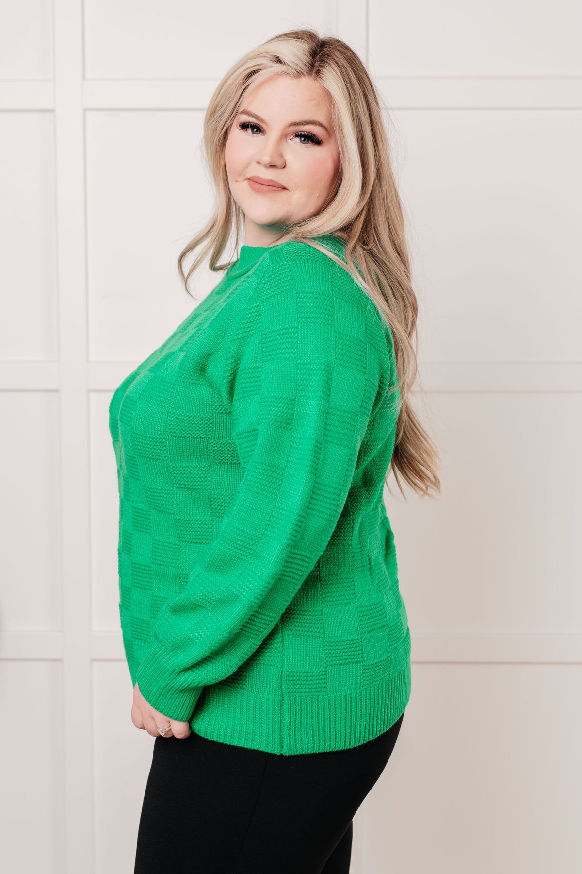 Woman modeling a green monochromatic checkered sweater knit pullover with ribbed trim and black pants against a paneled white wall