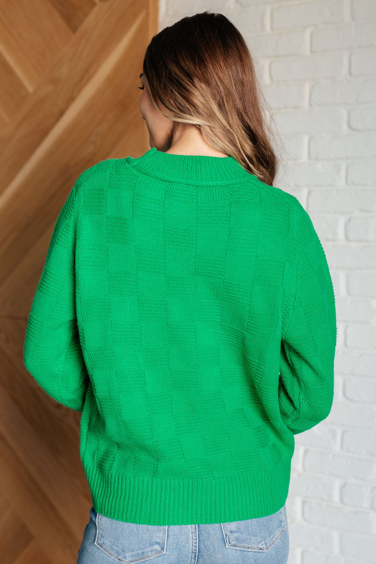 Back view of a woman wearing a green monochromatic checkered sweater knit pullover with ribbed trim and light wash jeans indoors