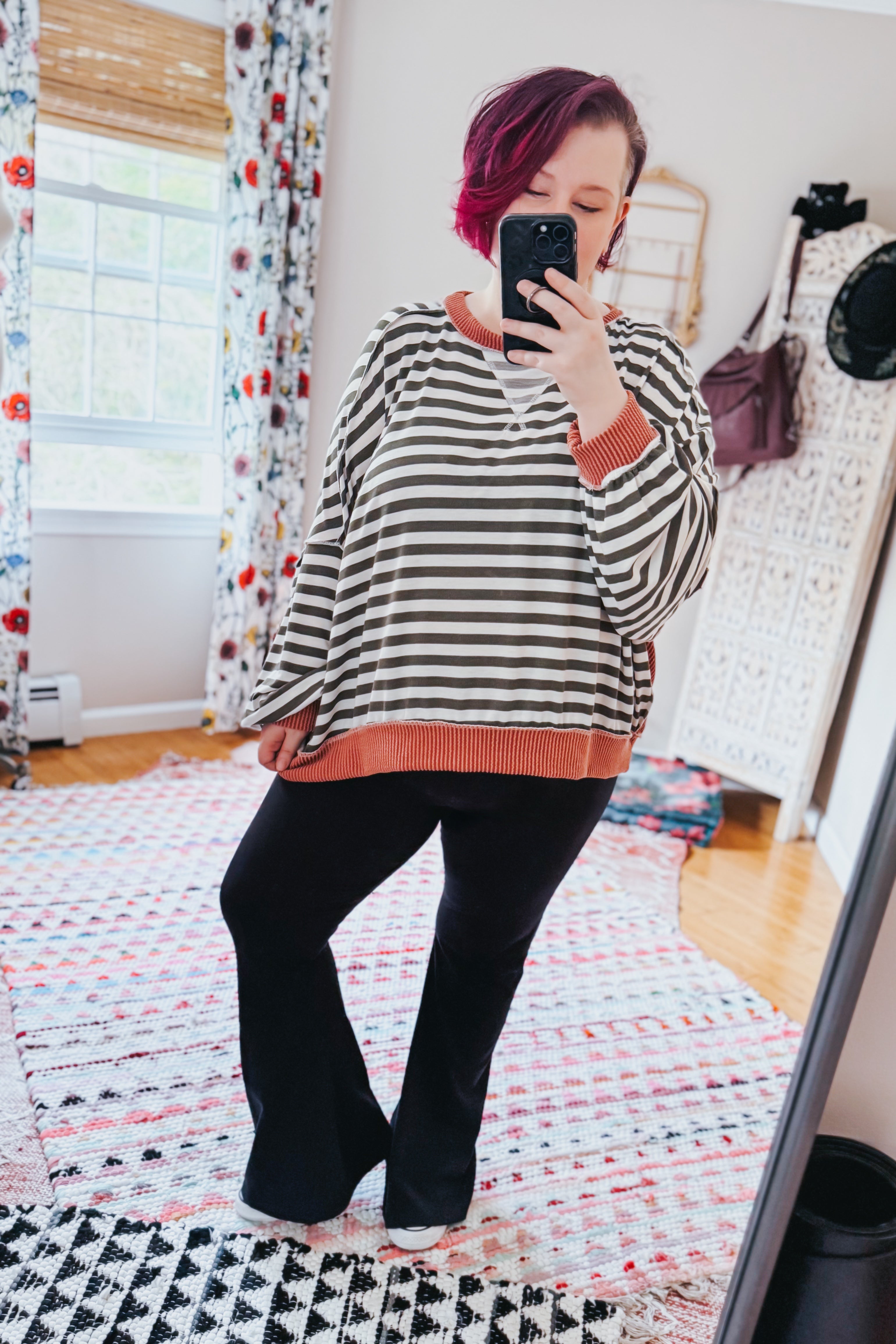 Woman with short magenta hair wearing striped pullover with rust trim and black pants taking mirror selfie indoors