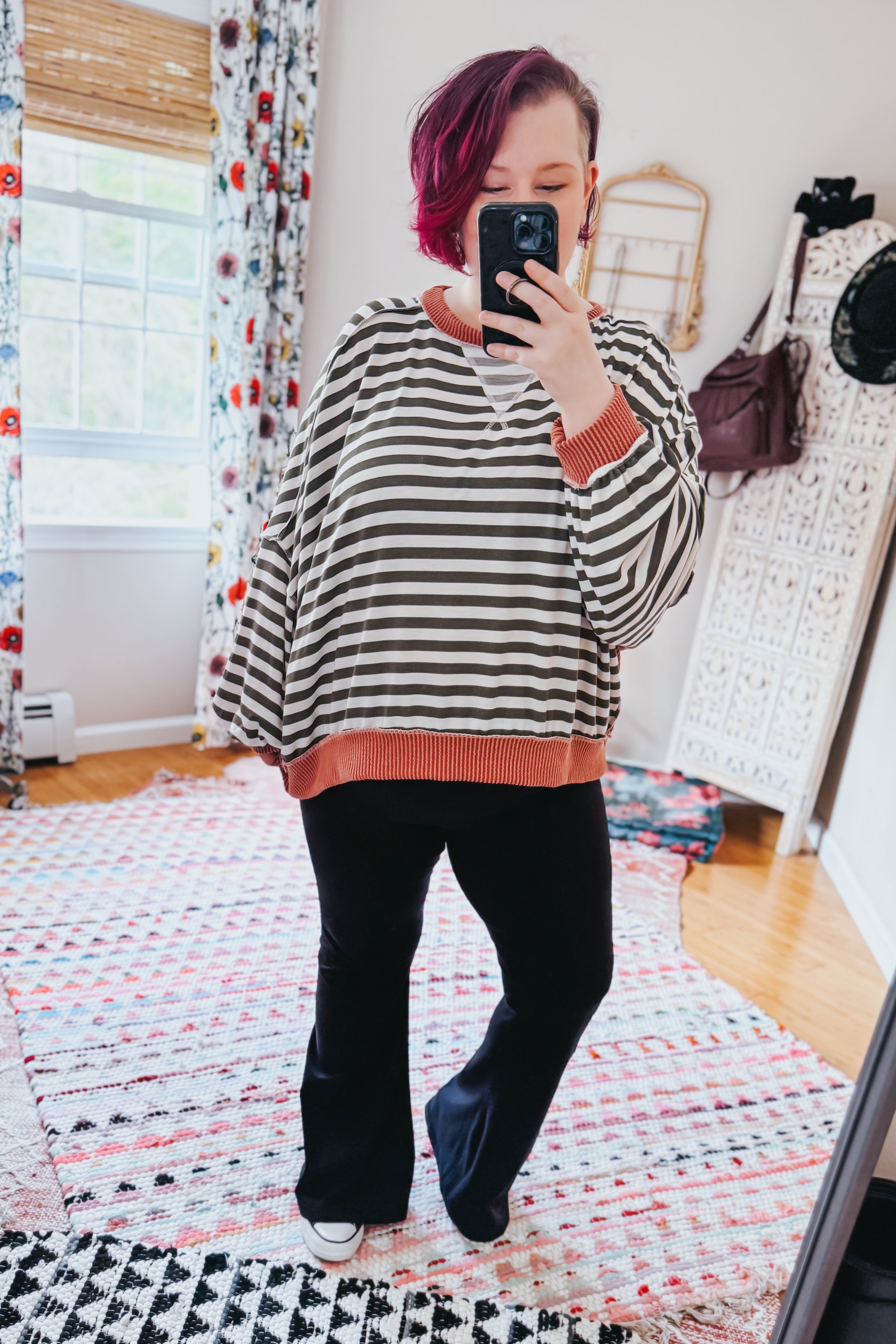 Woman with short pink hair taking mirror selfie wearing striped crew neck pullover with rust trim and black pants indoors