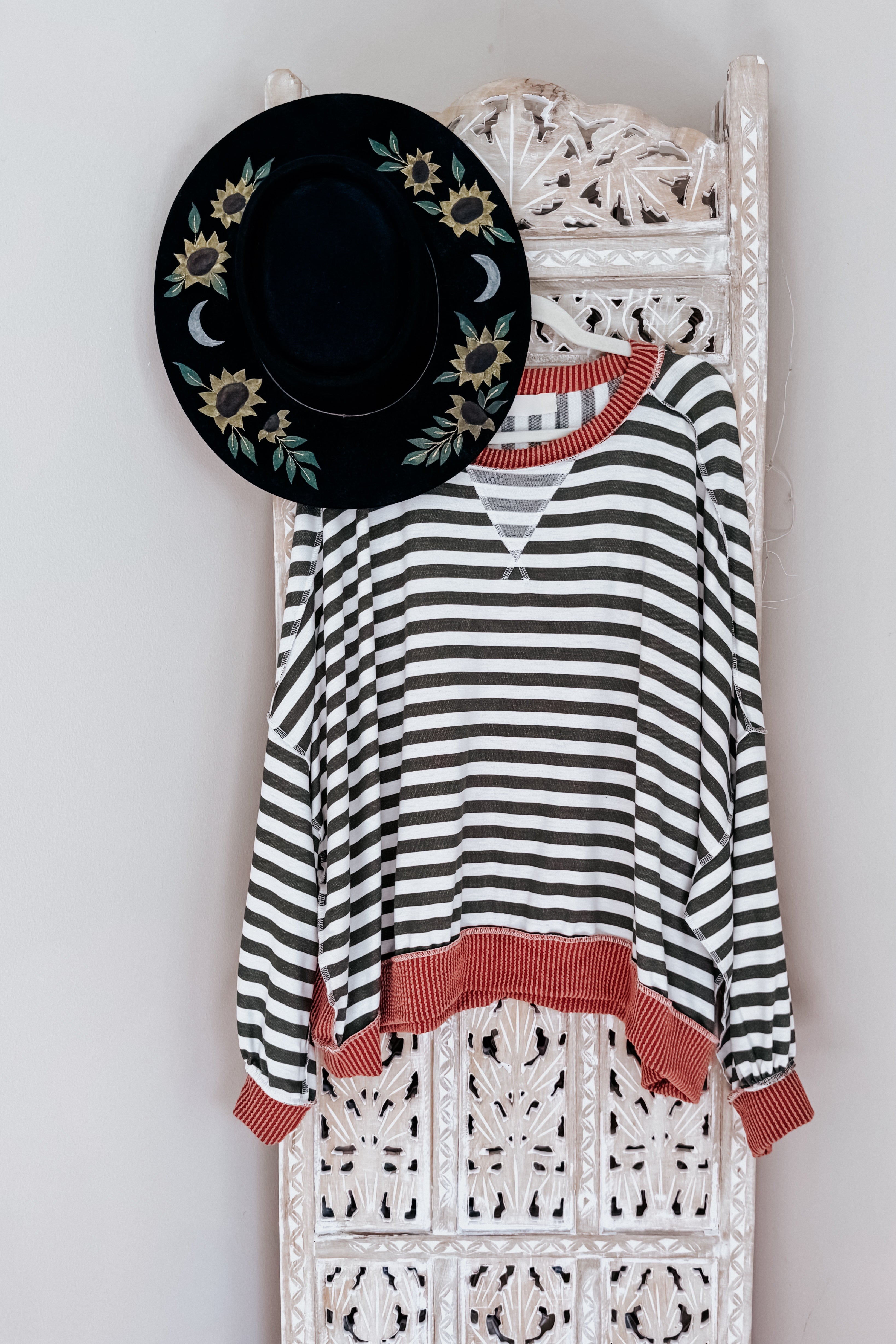 Striped crew neck pullover with rust trim hanging on carved white wooden screen with black embroidered sunflower hat