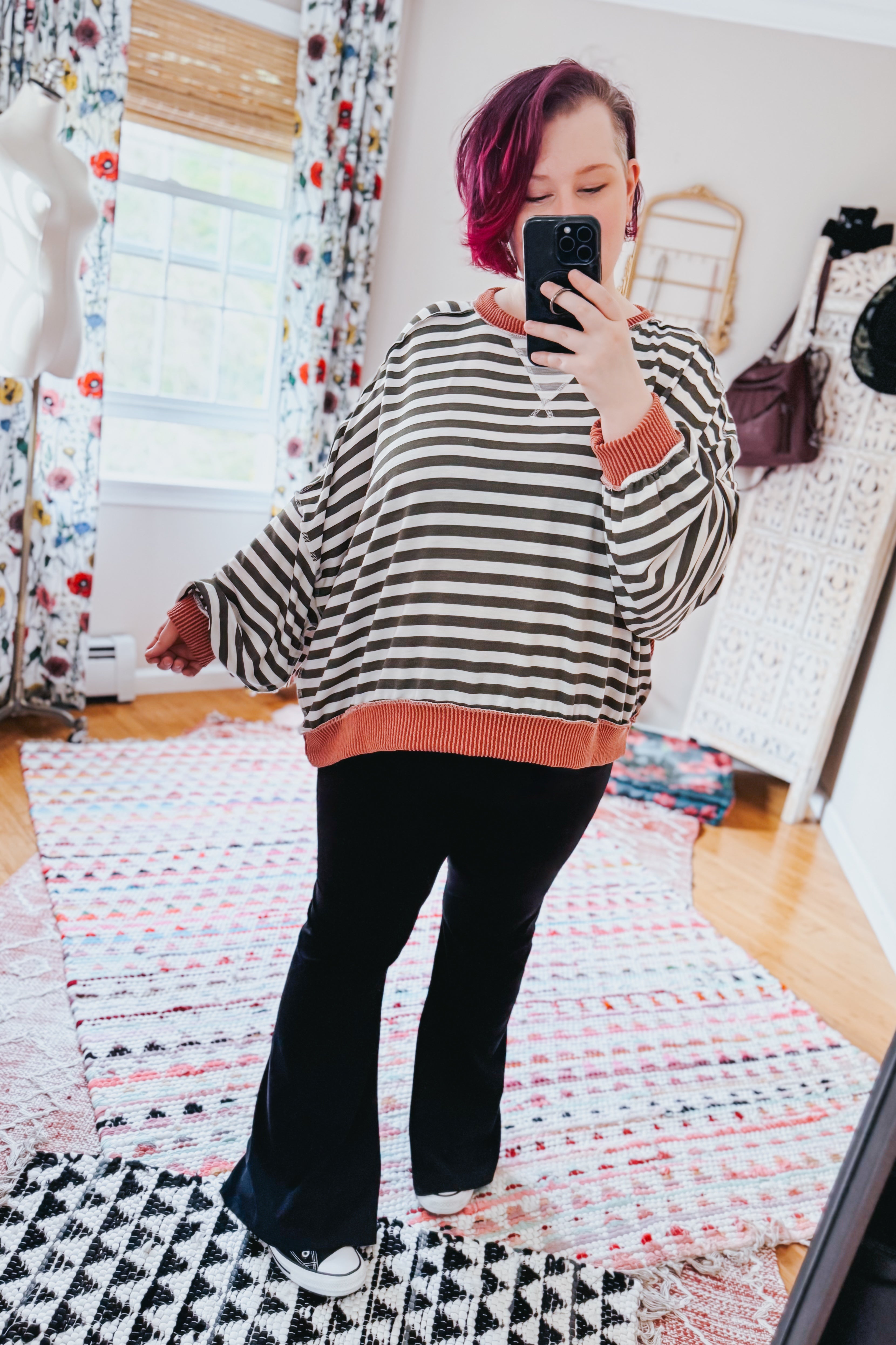 Woman with short magenta hair wearing striped crew neck pullover with rust trim taking mirror selfie indoors