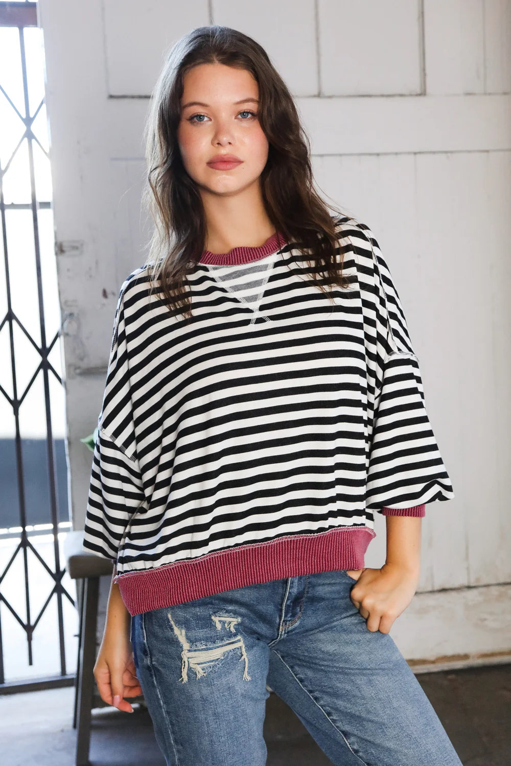 Woman wearing black and white striped pullover with wine ribbed neckline and hem standing indoors by window and door