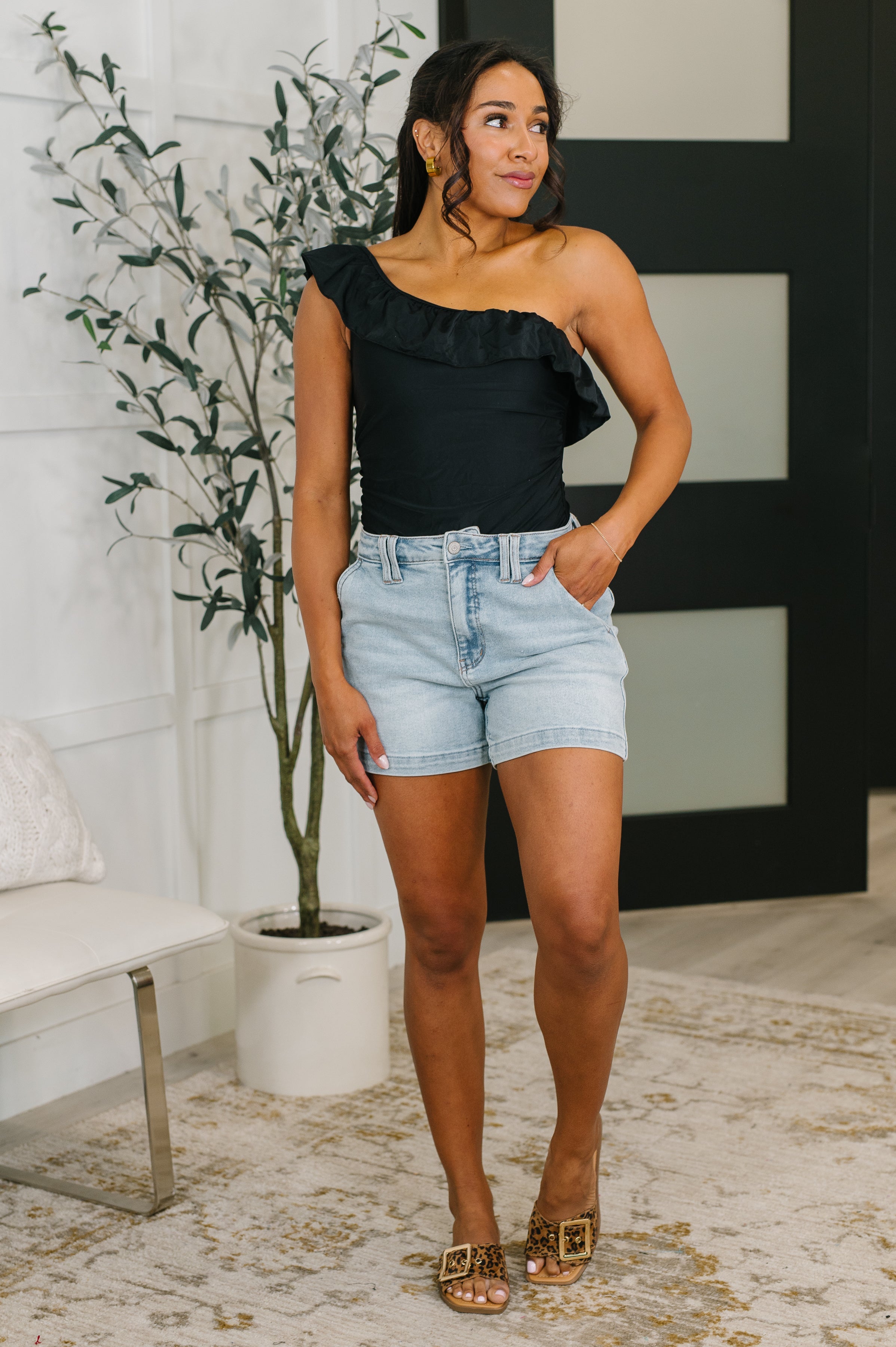 Woman wearing black one-shoulder top with ruffle detail, light denim shorts, and leopard print slide sandals indoors