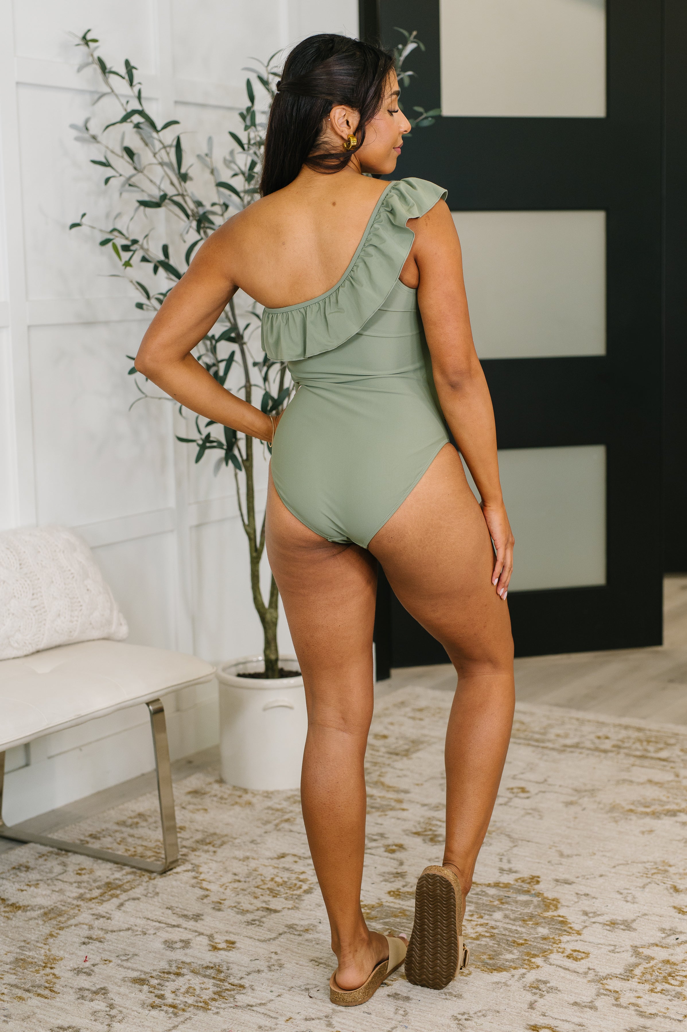Woman wearing olive one-shoulder one-piece swimsuit with ruffle detail, standing indoors on patterned rug