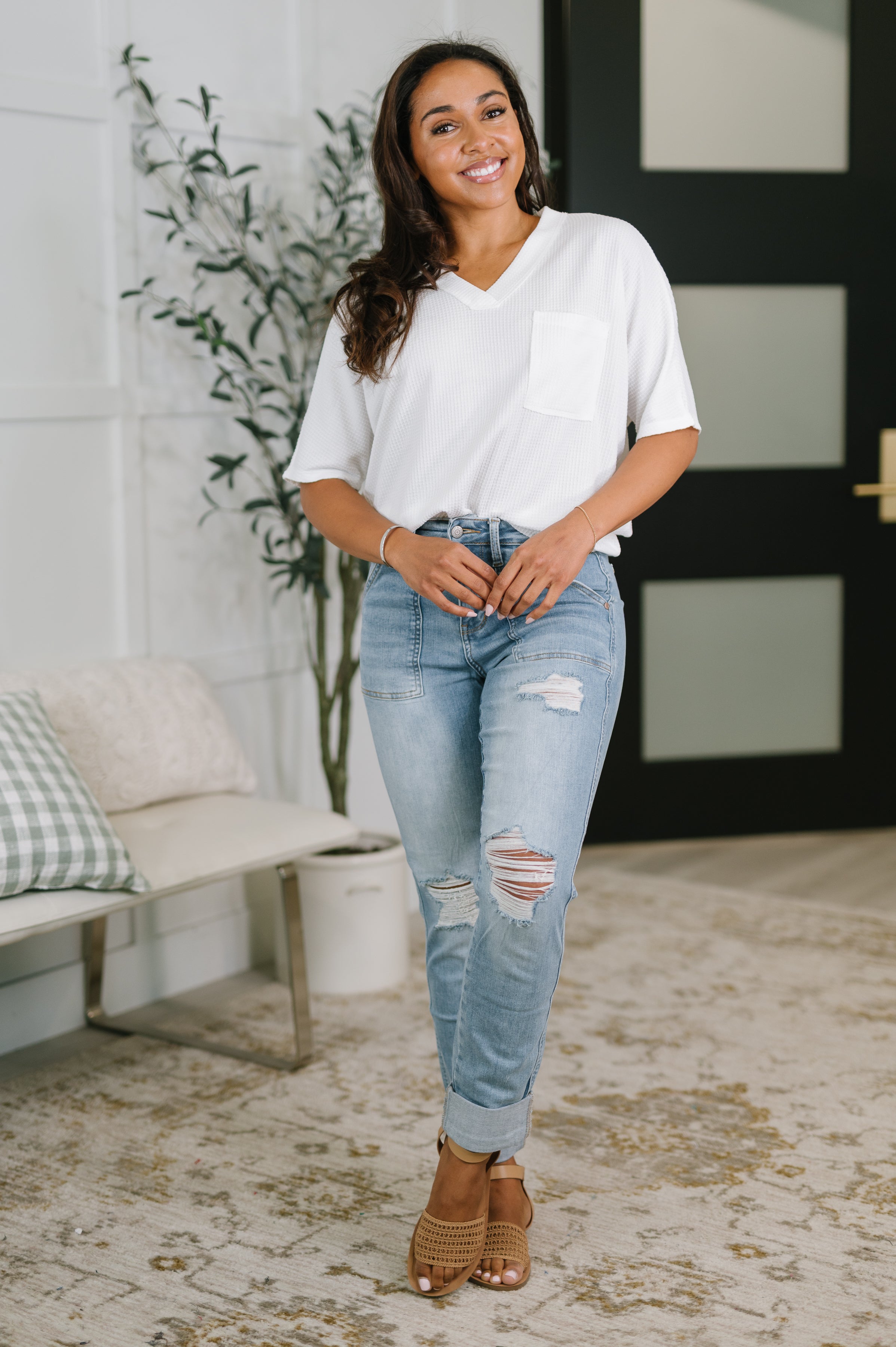 Smiling woman wearing light blue distressed boyfriend jeans and a loose white top in a cozy indoor setting
